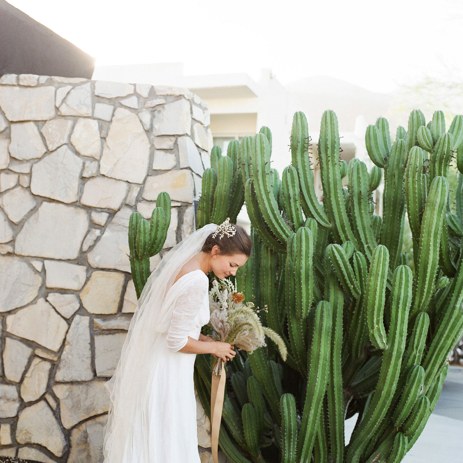 A medium shot of our bride from the side holding her bouquet and looking down towards her shoes standing in front of a huge cactus and natural brick wall. 