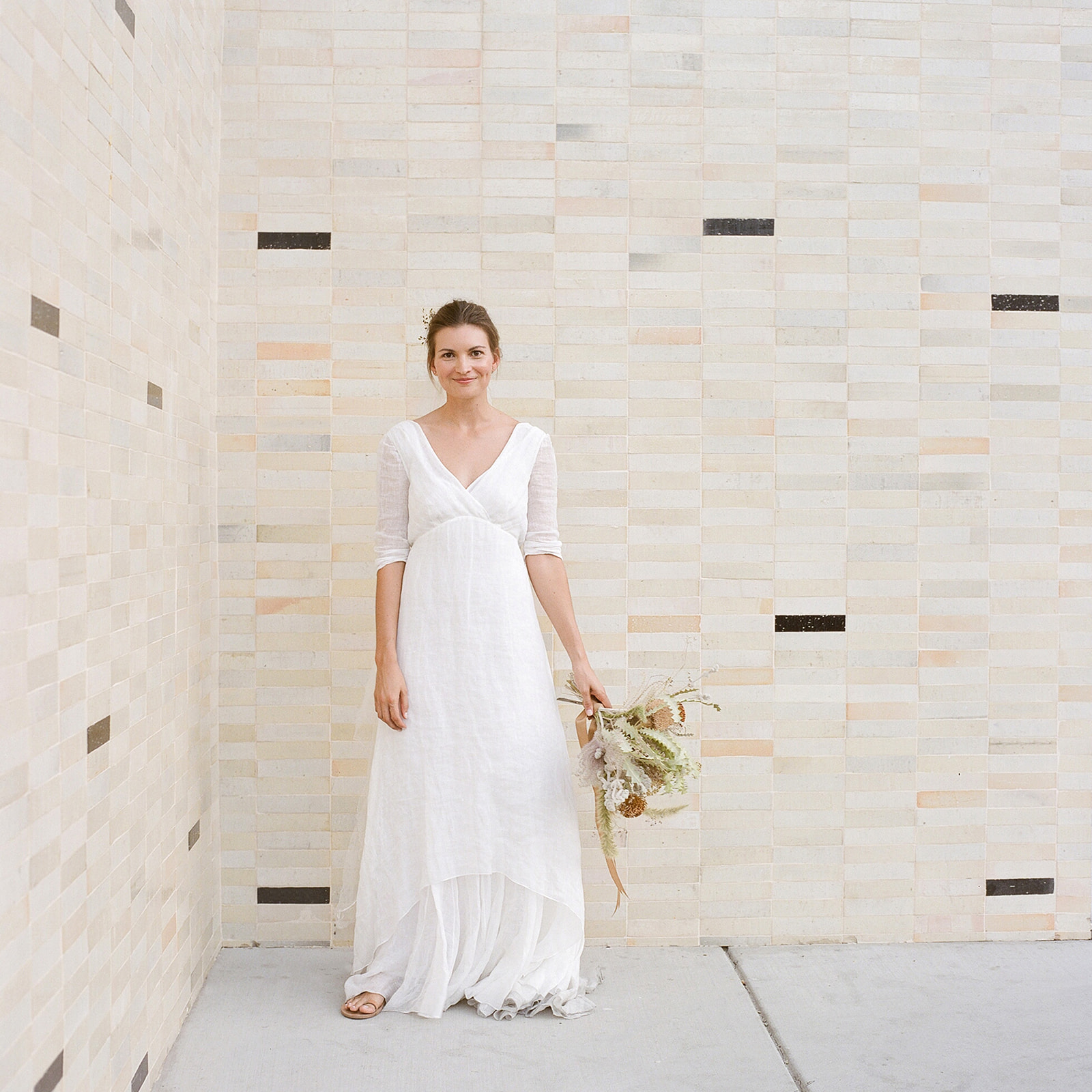 Wide shot of our cheerful and happy looking bride K holding her bridal bouquet in her left hand. She wears a gauzy floor length off white gown with simple flat tan sandals.  