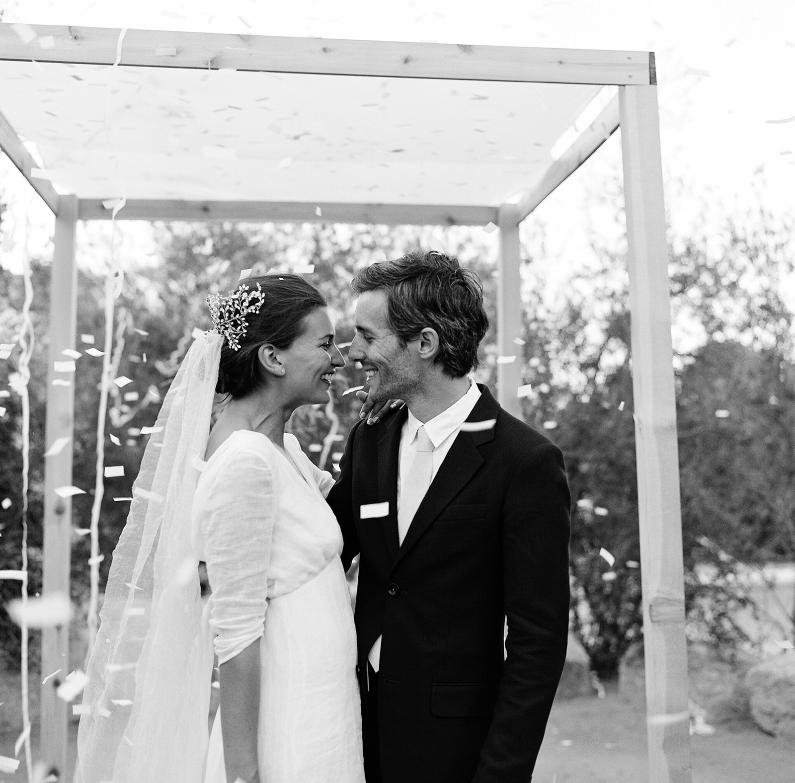 Black and white medium shot of K and E smiling under the Chuppah they made themselves. Confetti is flying in the air!