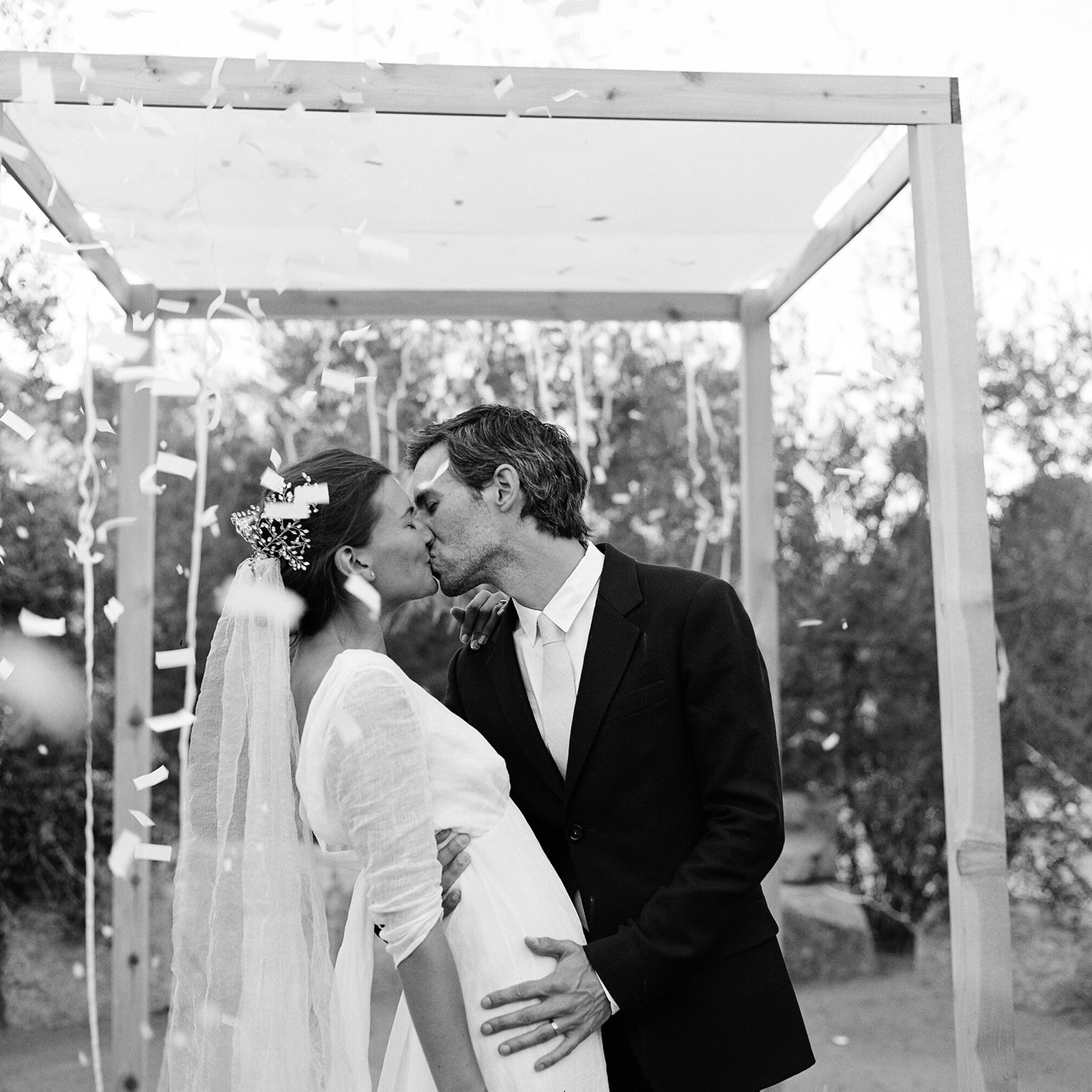 Black and white medium shot of K and E kissing under the Chuppah they made themselves. Confetti is flying in the air!