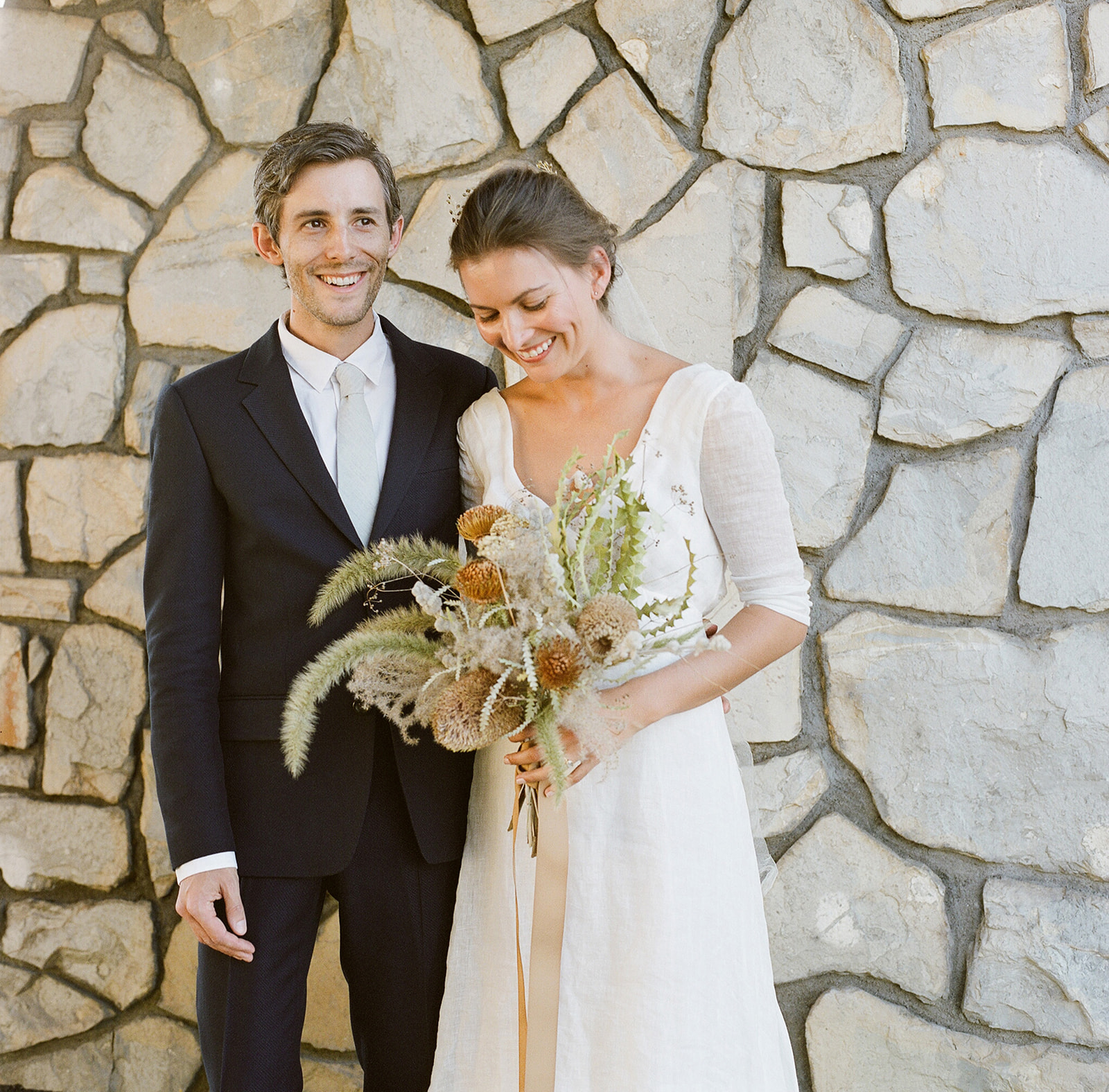 Medium shot of K and E smiling andlooking away from the camera standing in front of a natural brick wall. 