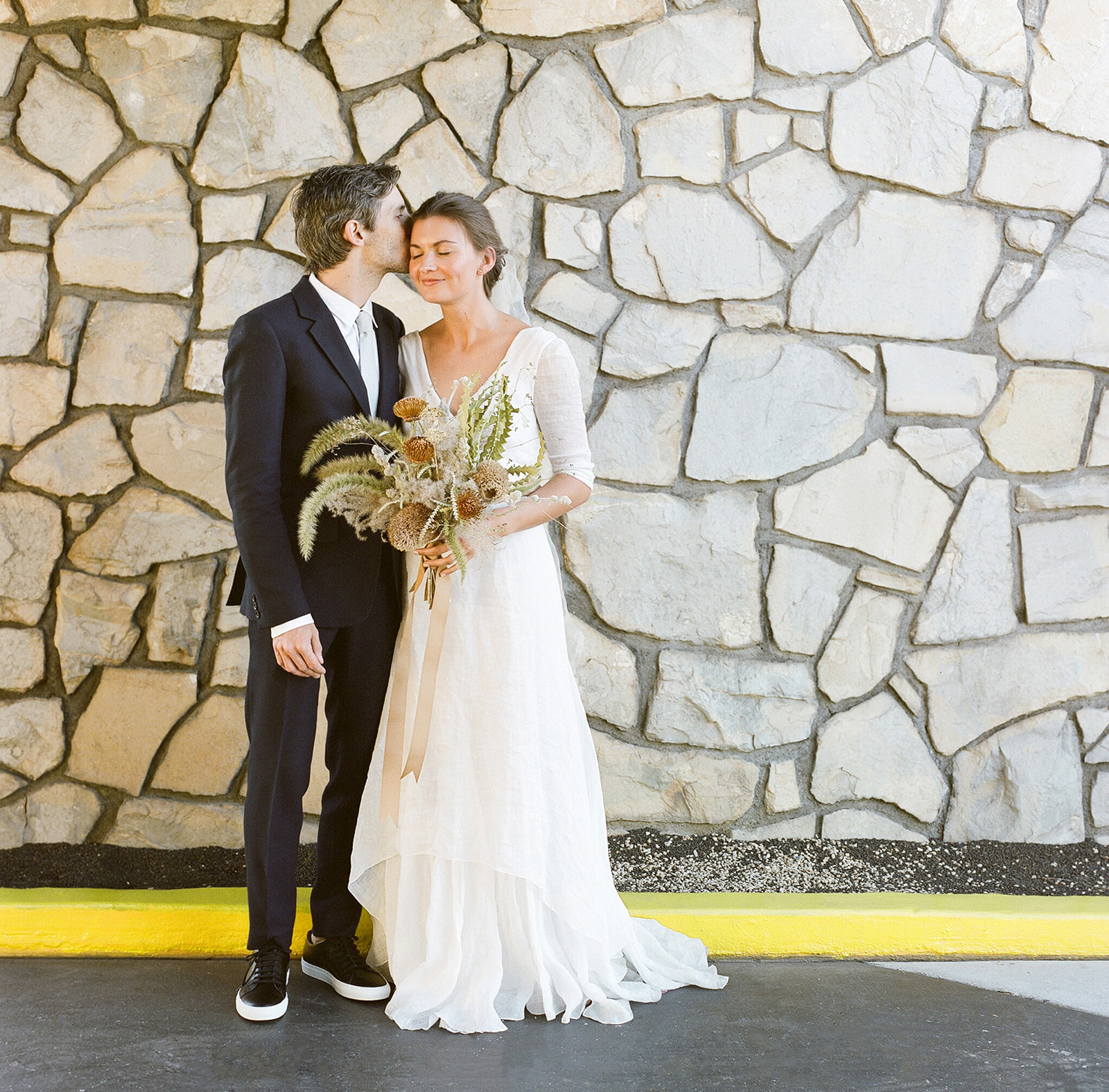 Wide shot of the couple standing in front of a natural brick wall with a bright yellow curb at their feet. He kissing her head while her eyes are closed. 