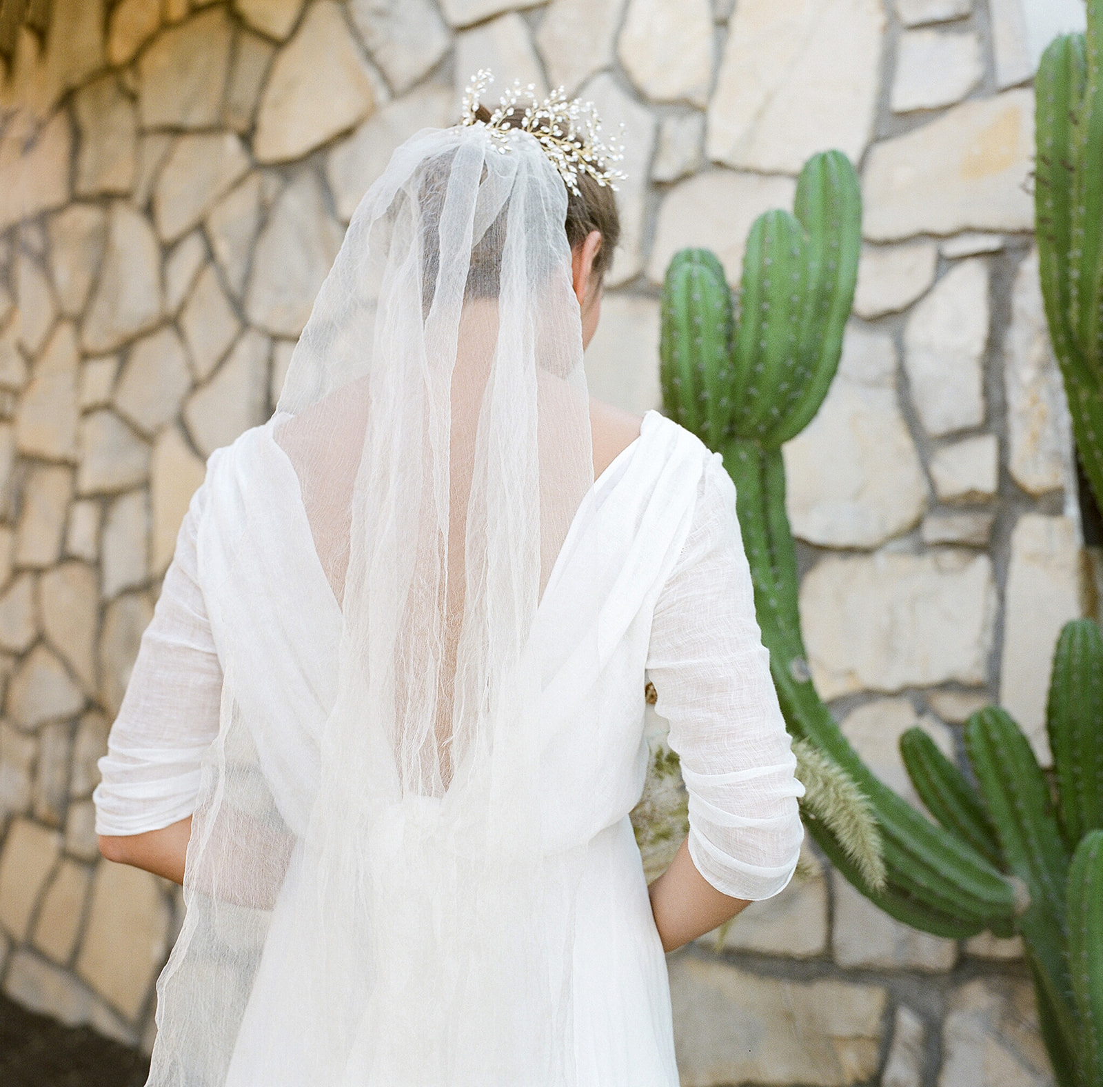 A medium shot of our bride with her back to camera. She wears a light gauzy veil that sits on the back of her hair that is pulled back in a bun. She stands in front of cactus and natural brick wall. 