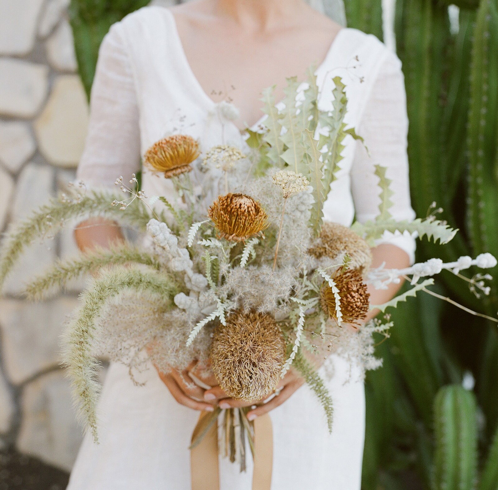 Close up shot of the brides hands holding a wild California native bouquet designed by the bride featured creosote, white sage, buckwheat and with banksia as the focal point. 