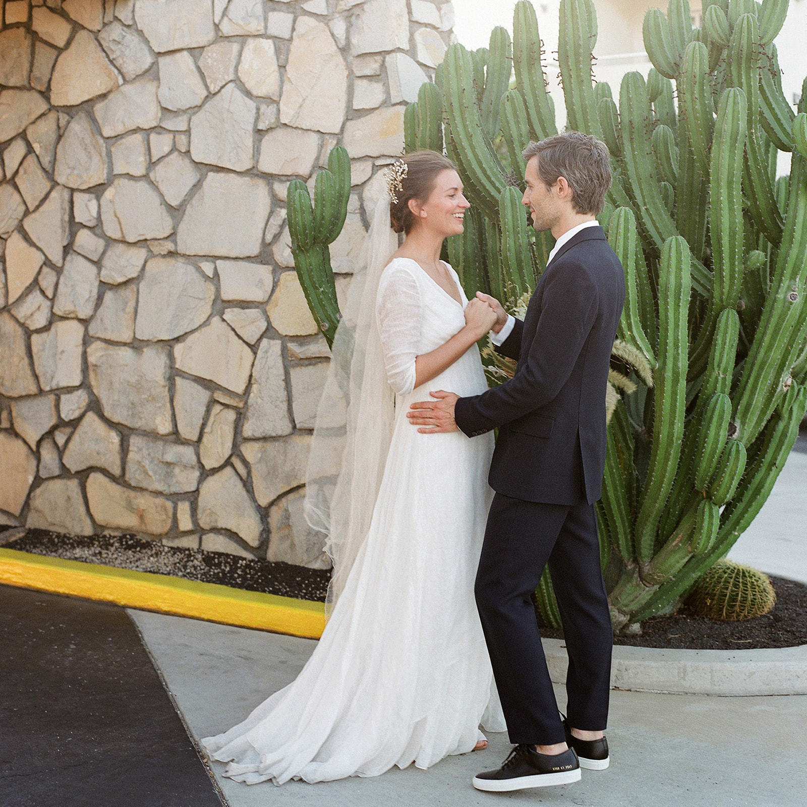Wide shot of the couple standing in front of a large cactus and a natural brick wall with a bright yellow curb at their feet.