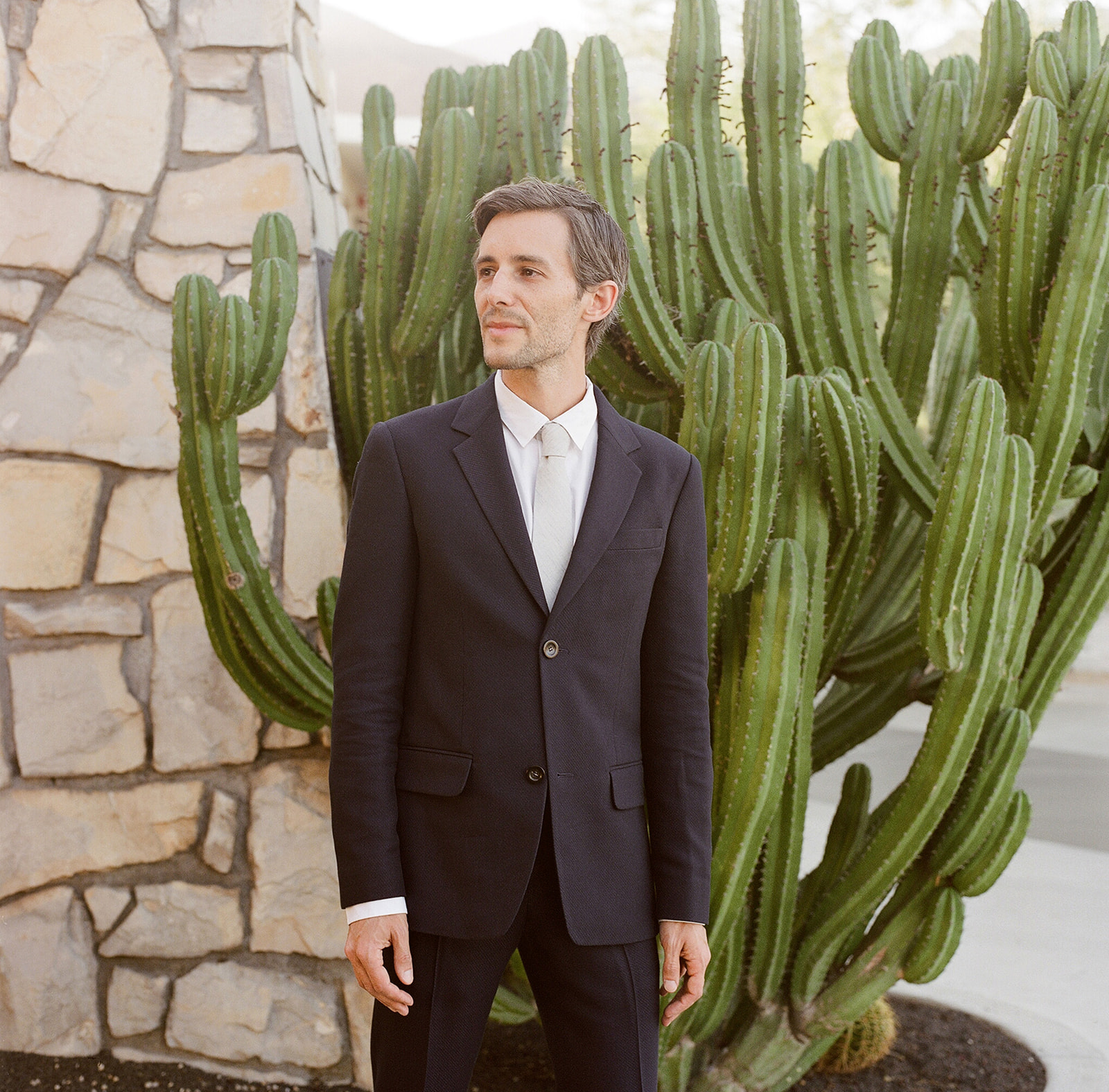 Medium shot of our groom E in his black tux with white button up shirt and gray tie standing in front of a huge cactus and natural brick wall. 