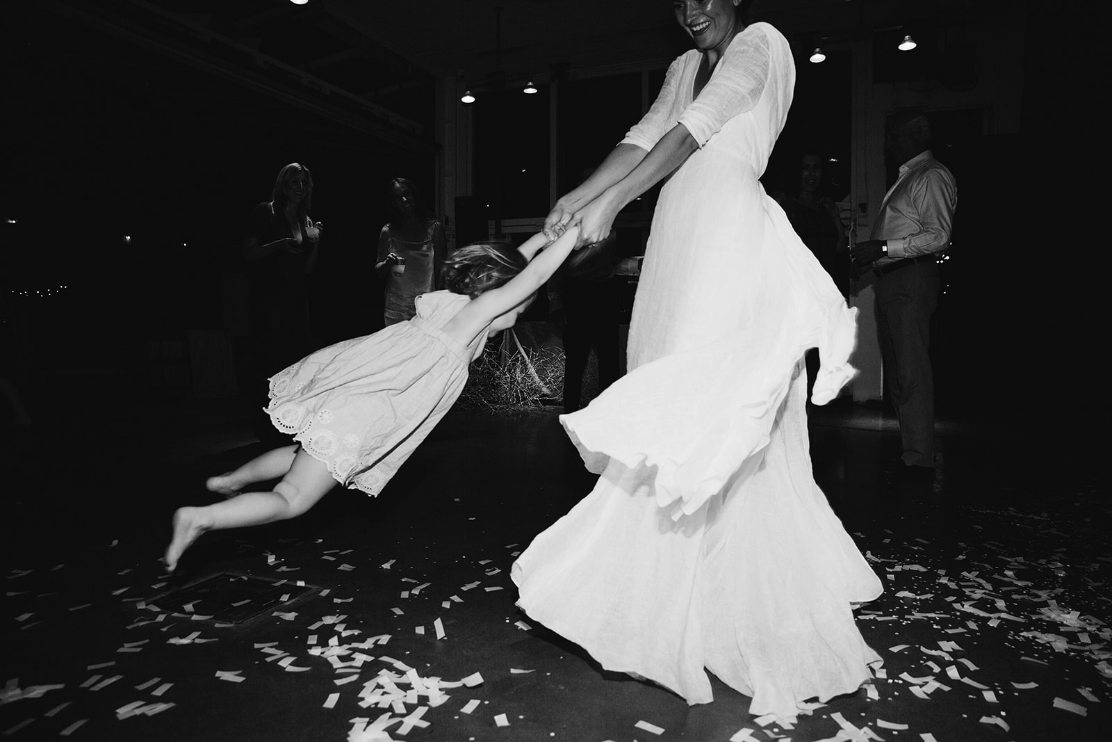 Black and white candid shot of K spinning a child around in a circle on the dance floor that is covered in white paper confetti. 