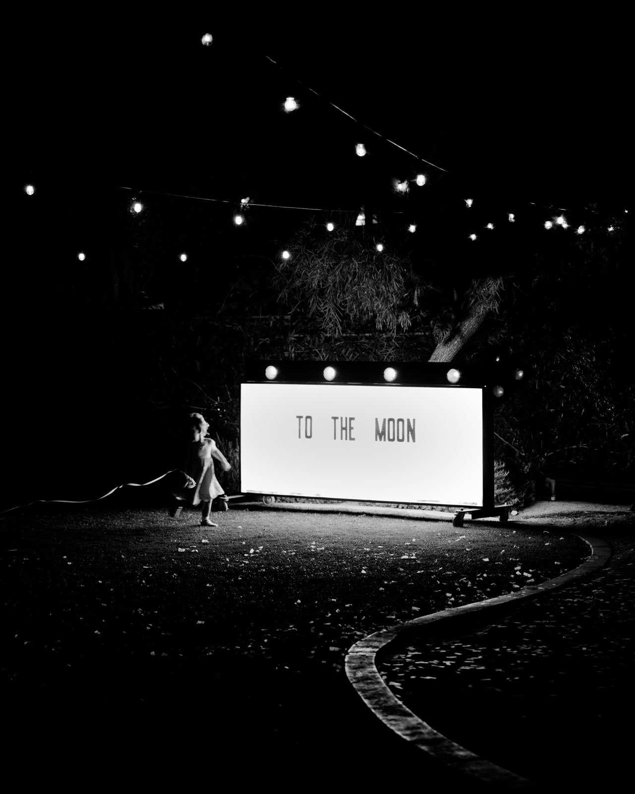 Black and white candid of a large marquee sign that is very bright stating the words "to the moon" while a little girl runs by .
