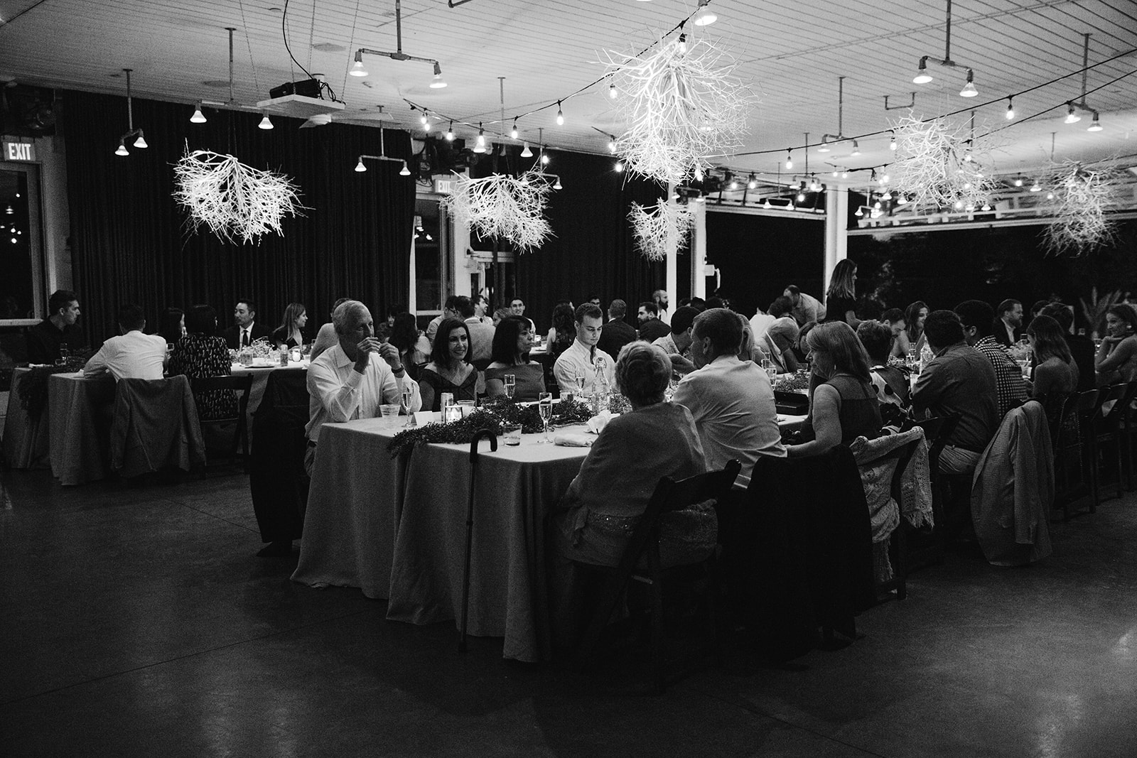 A black and white extra wide shot of one of the long tables with guests at the tables with tumbleweeds above their heads. 