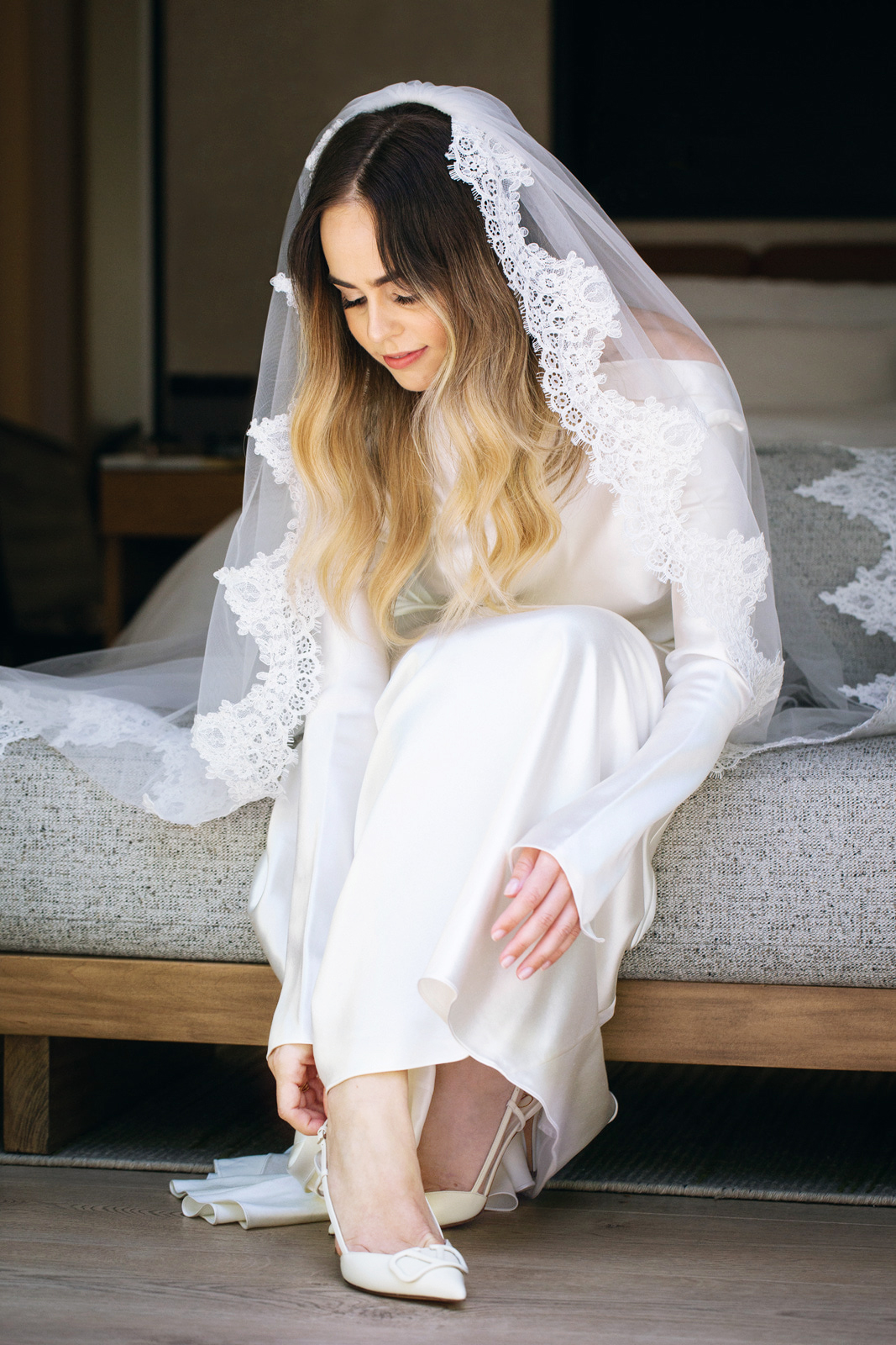 Wide shot of M sitting on the end of a bed in her white bridal gown and veil that sits at the back of her head. She is putting her right shoe on and is almost ready to go to the church.
