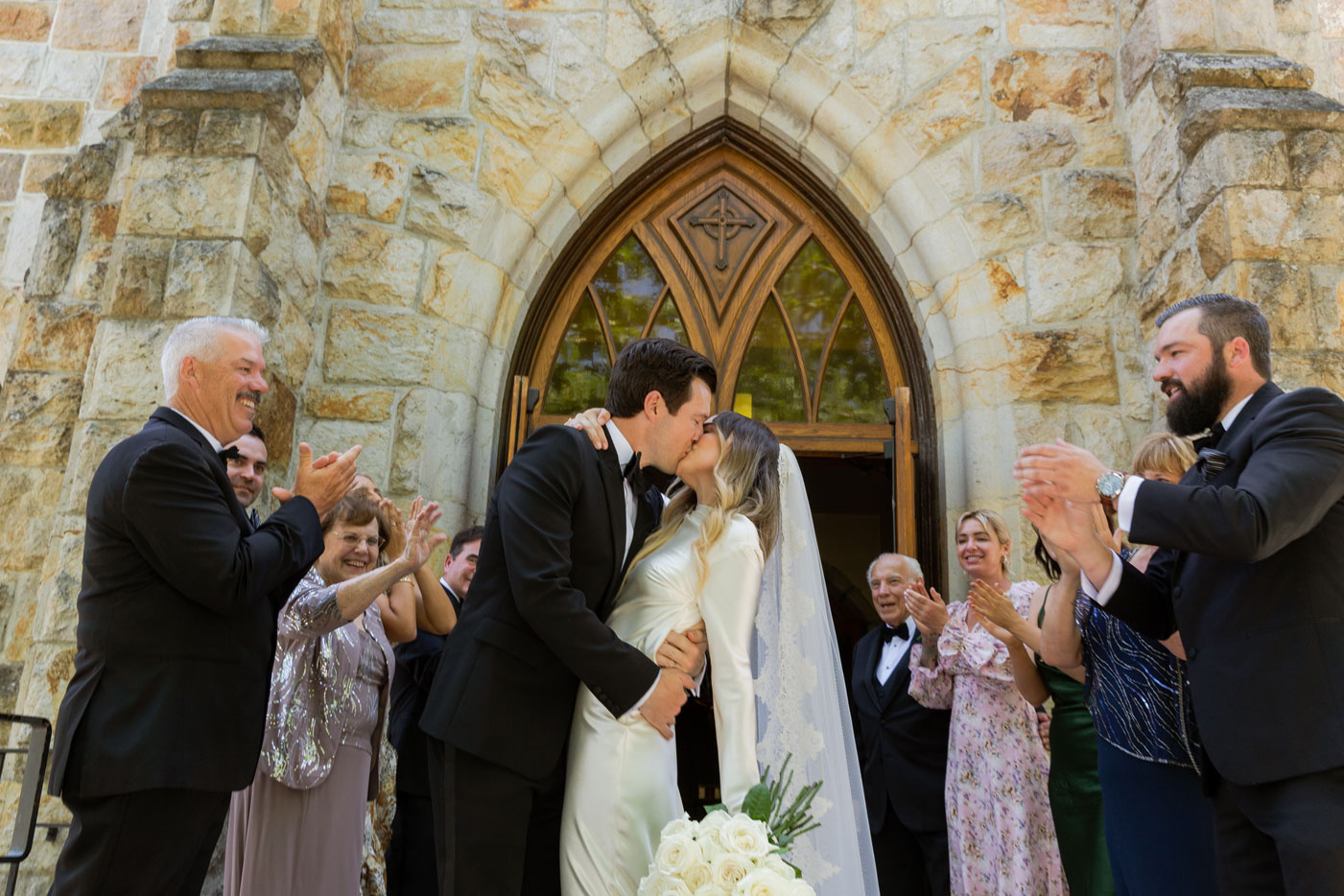 Wide shot of M and C kissing in front of the church with their friends and family all around them clapping and smiling.