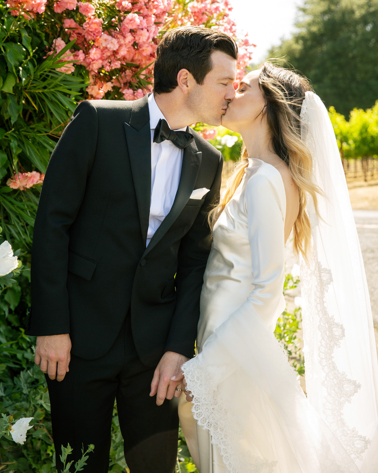 Medium shot of M and C kissing in front of green foliage and pink flowers. The sun shines through M’s veil and you can see grape fields in back of them.