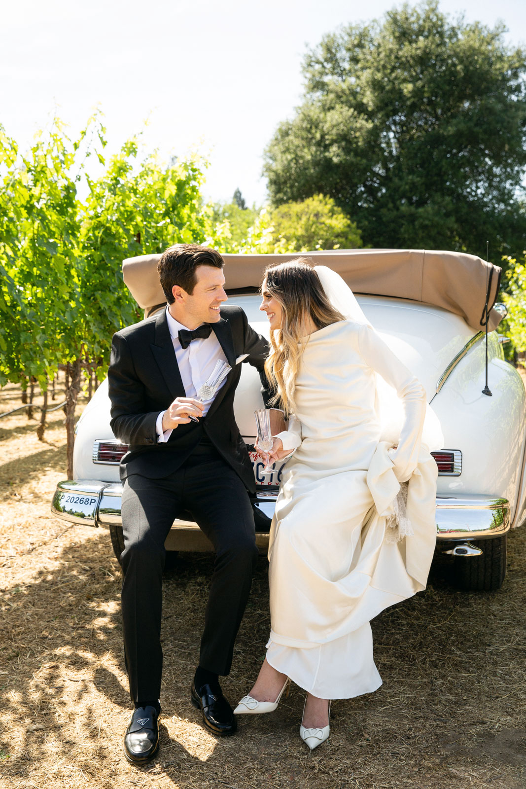 Wide shot of the couple sitting and talking on the back of a white vintage car with glasses of champagne in their hands.