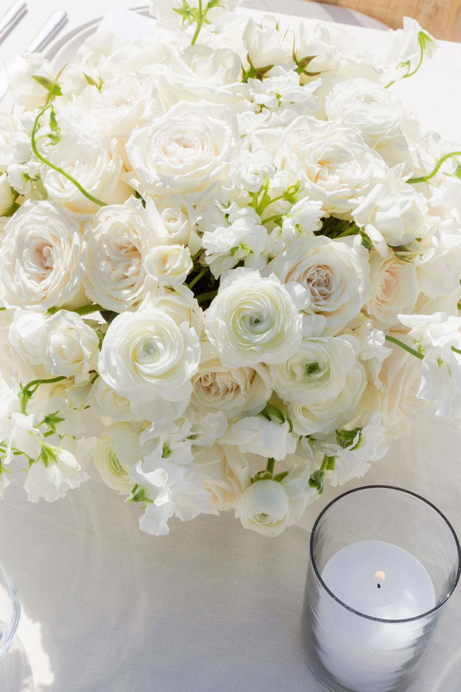Close up shot of an all white centerpiece besides a lit candle votive..
