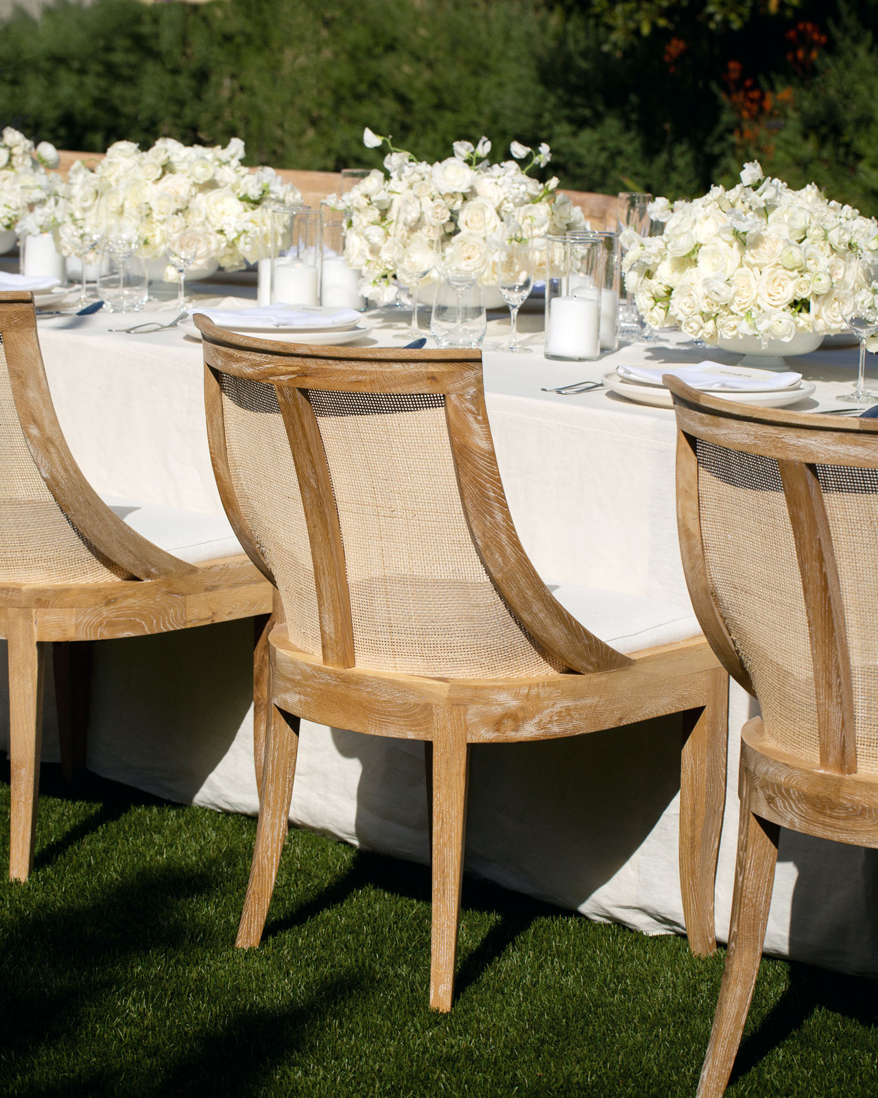 Medium shot of the backs of three wooden chairs that sit in front of long white tablecloths and white centerpieces.