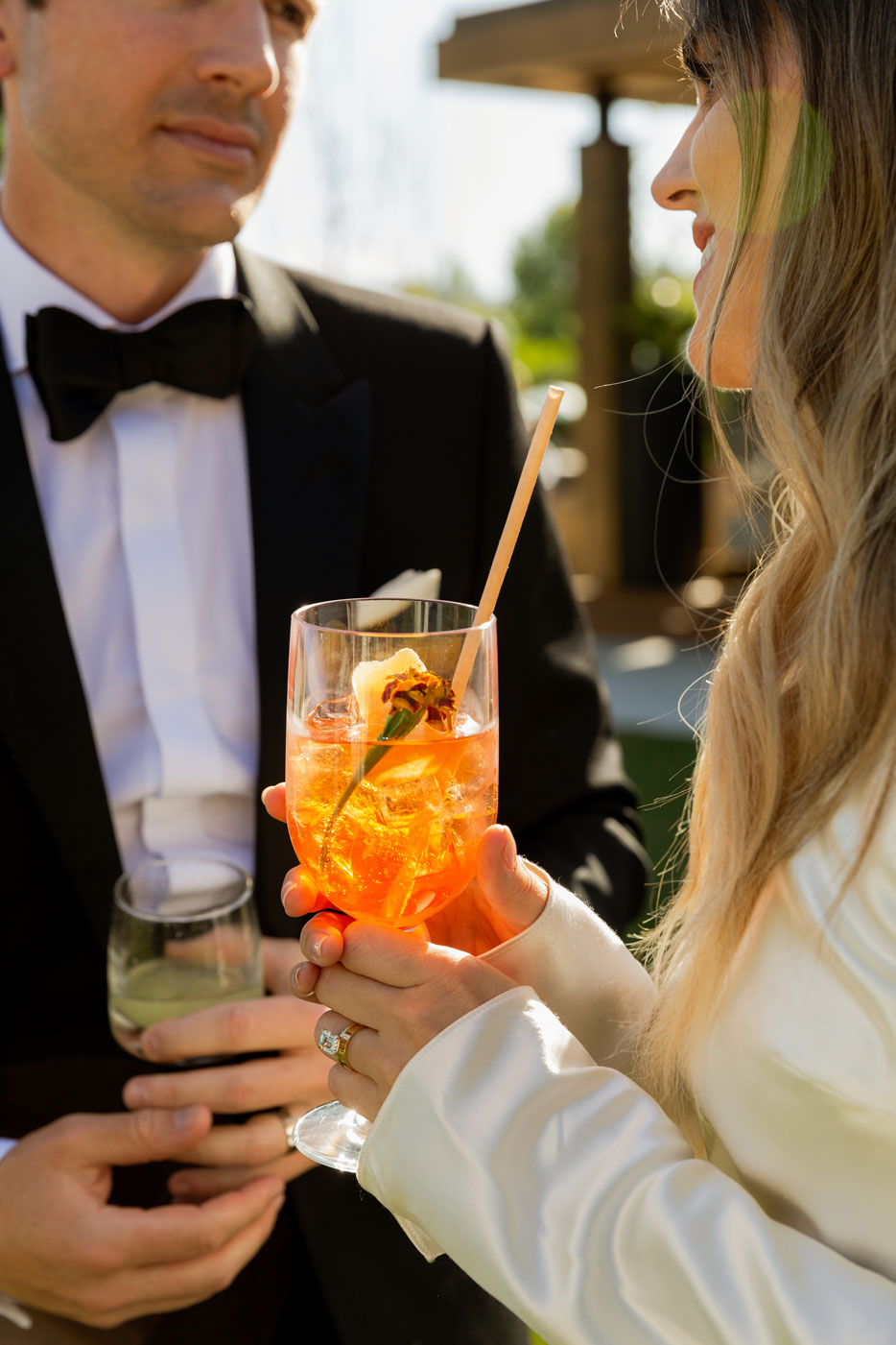 Close up shot of the couples hands enjoying their cocktails. M with a large goblet of an Aperol spritz that contains a small carnation flower. While C drinks from a low ball glass containing a cucumber.