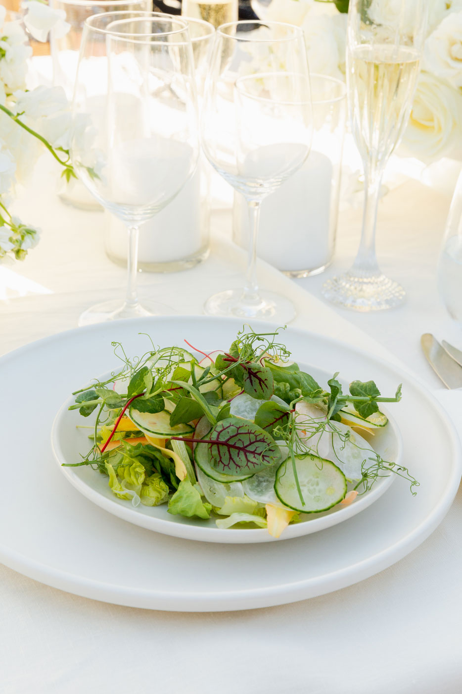 Close up shot of a garden salad on a white plate on a white charger sitting 0on a white tablecloth with glassware and unlit white candles.