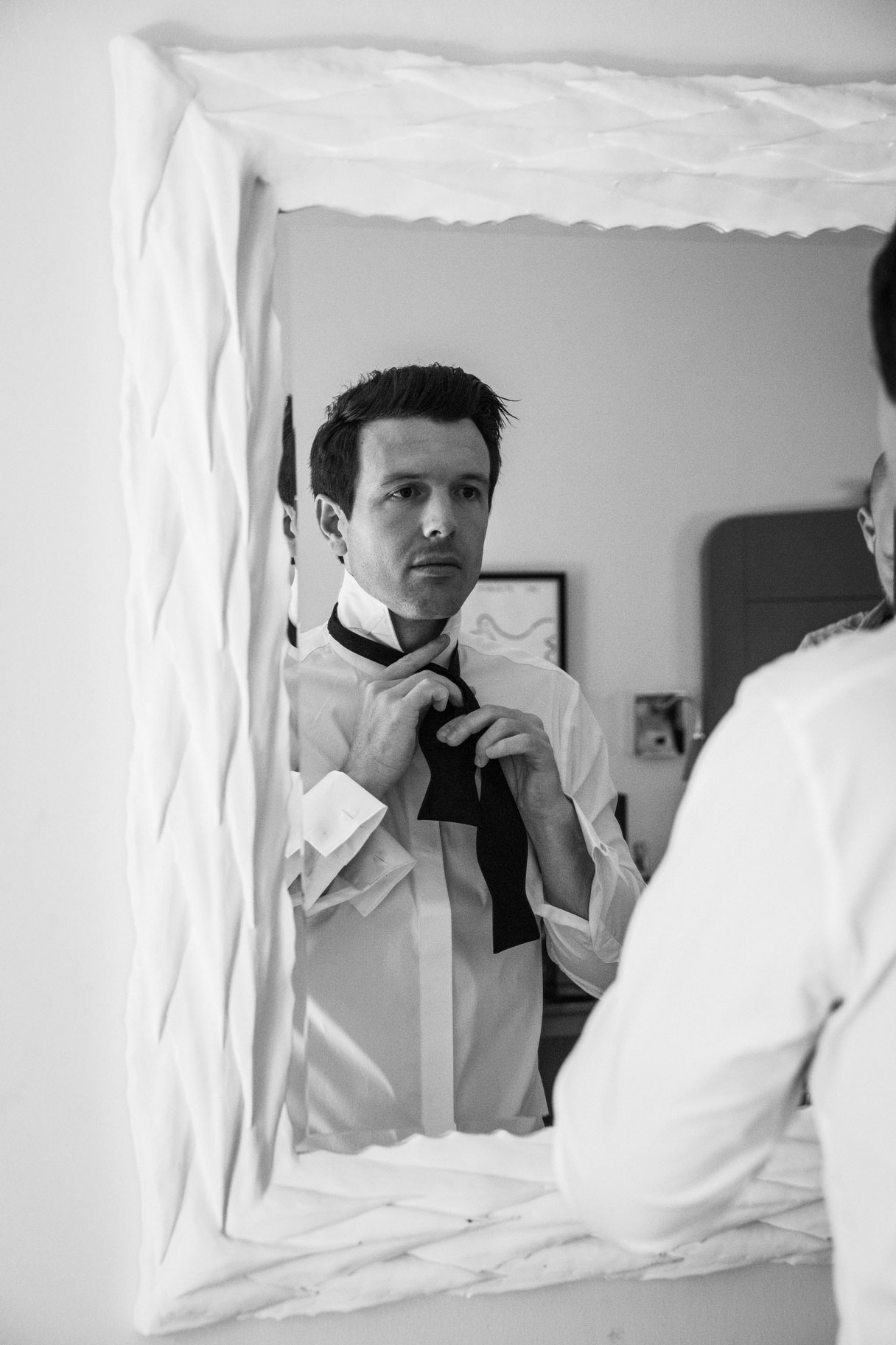 Black and white medium shot of our groom C wearing a white button-up shirt and looking in a large white framed mirror adjusting his black bow tie.