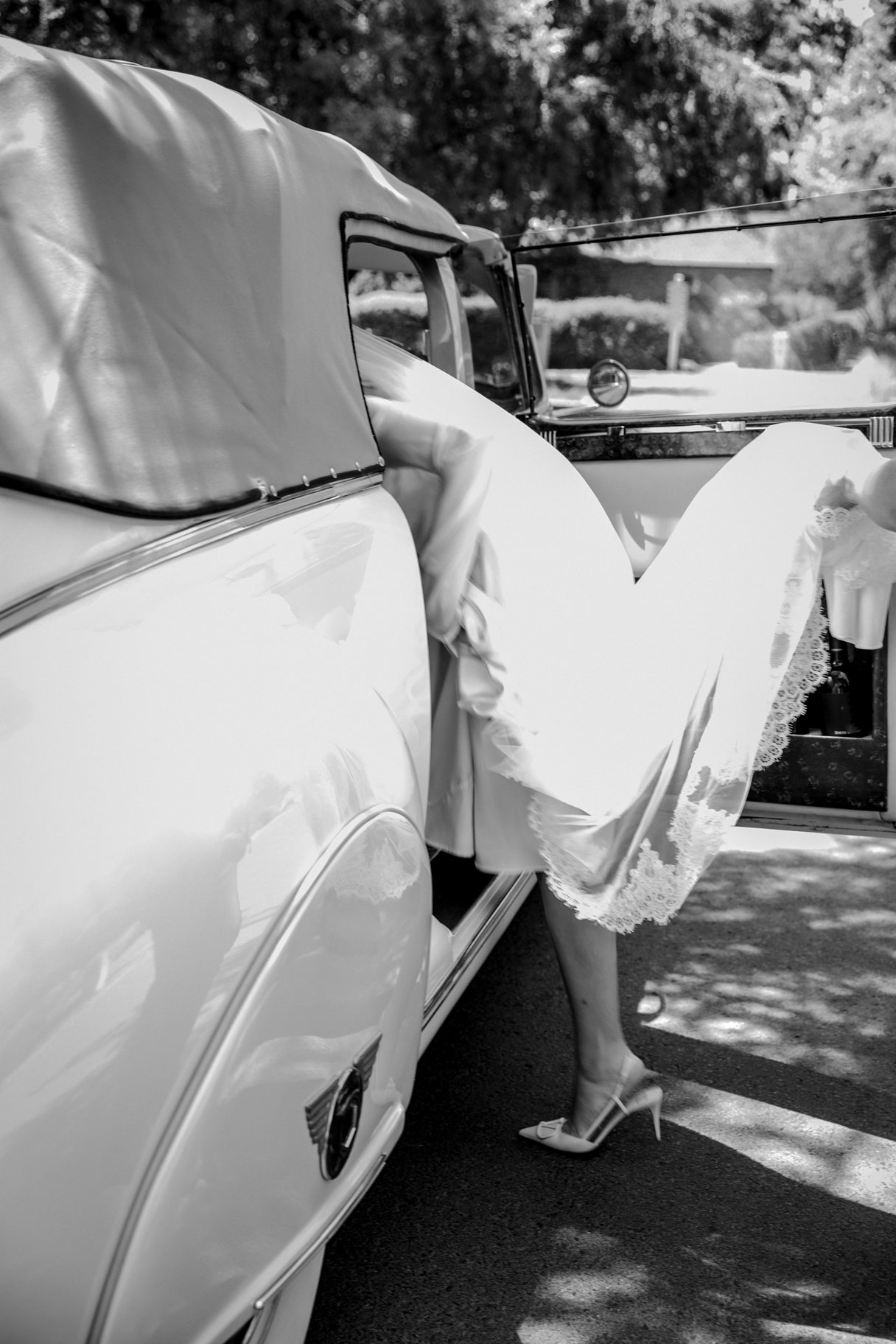 Black and white wide shot of M getting into a vintage car. Her head and half of her body are already inside the car, so the perspective is of her from behind.