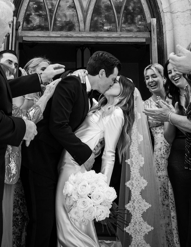 Black and white medium shot of M and C kissing in front of the church with their friends and family all around them.