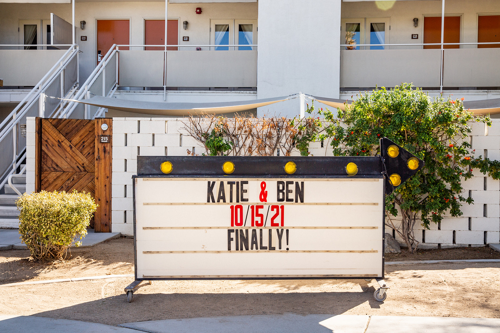 Wide shot of a billboard sign sitting on ground in front of hotel room. The sign reads "K and B 10/15/21 Finally!" The sign is a reference to the postponement of their wedding date twice due to the Covid 19 pandemic.