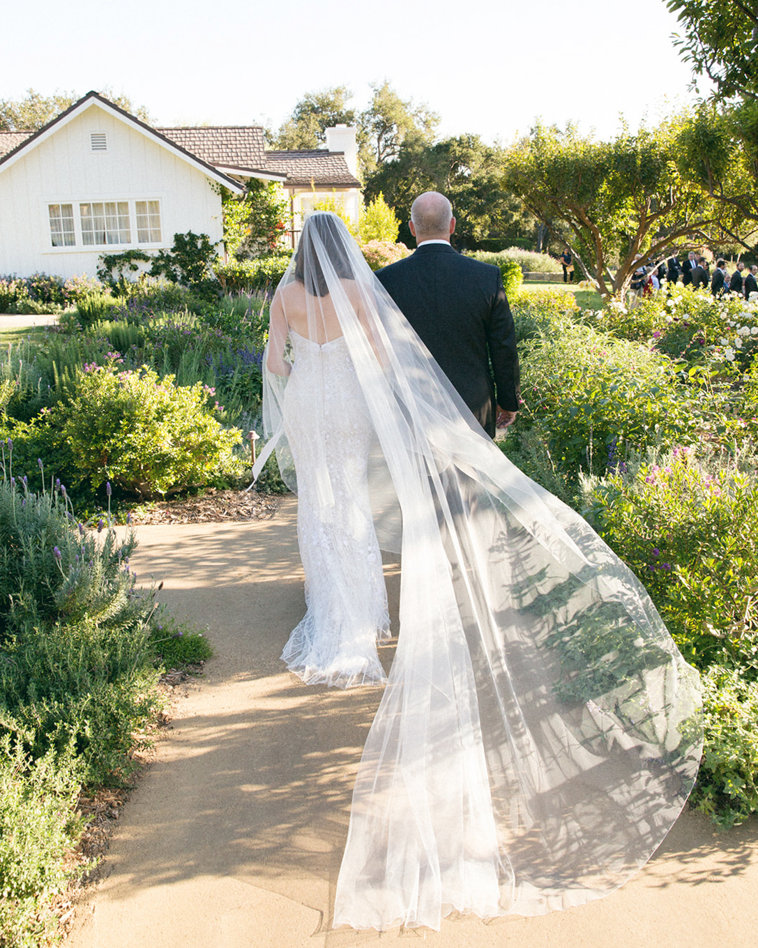 Wide shot of the back of the bride and her father walking down a garden path. Her veil is very large and epic and is so long it is touching the ground.
