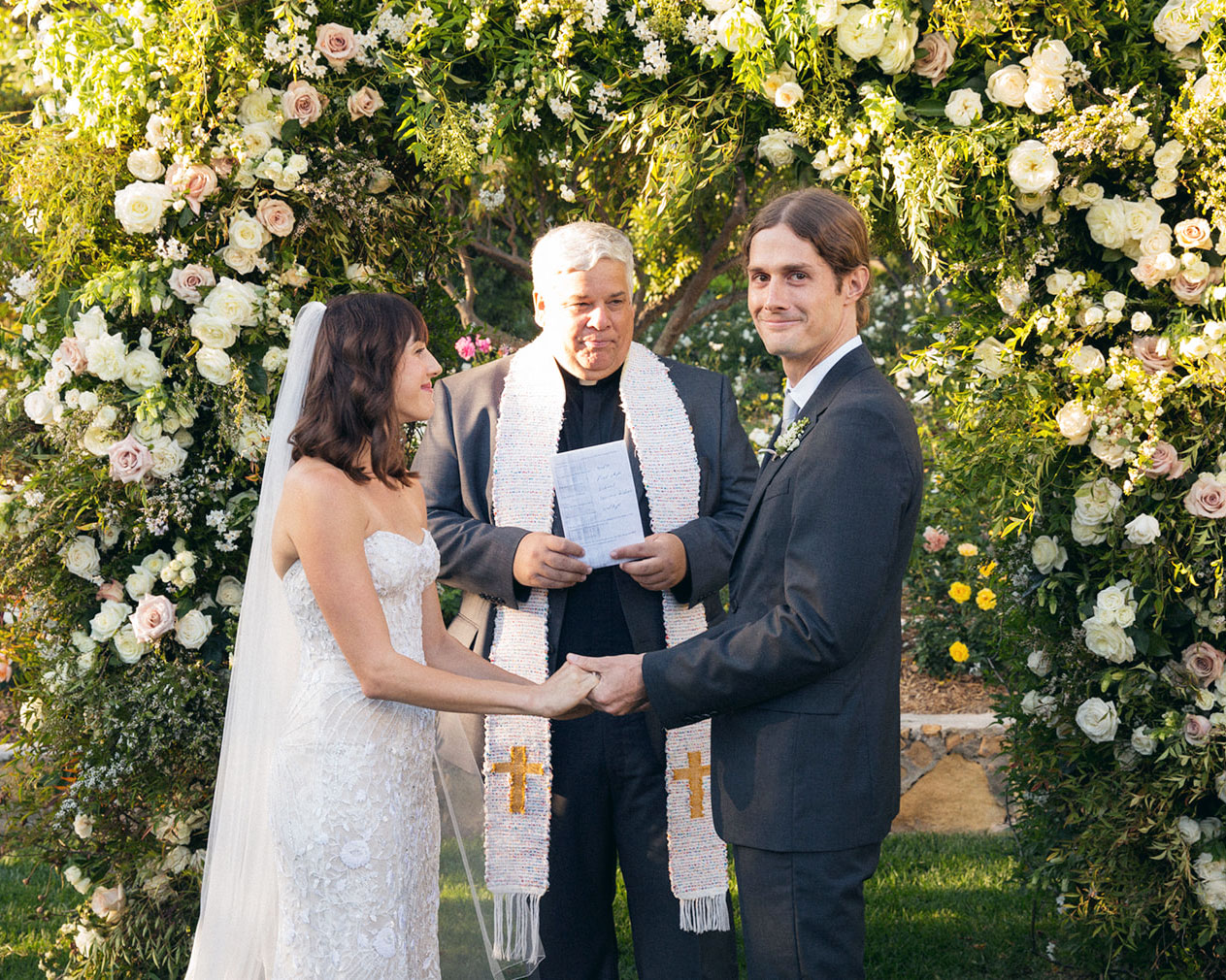 Medium wide shot of the bride, groom and pastor standing under a flower arch.