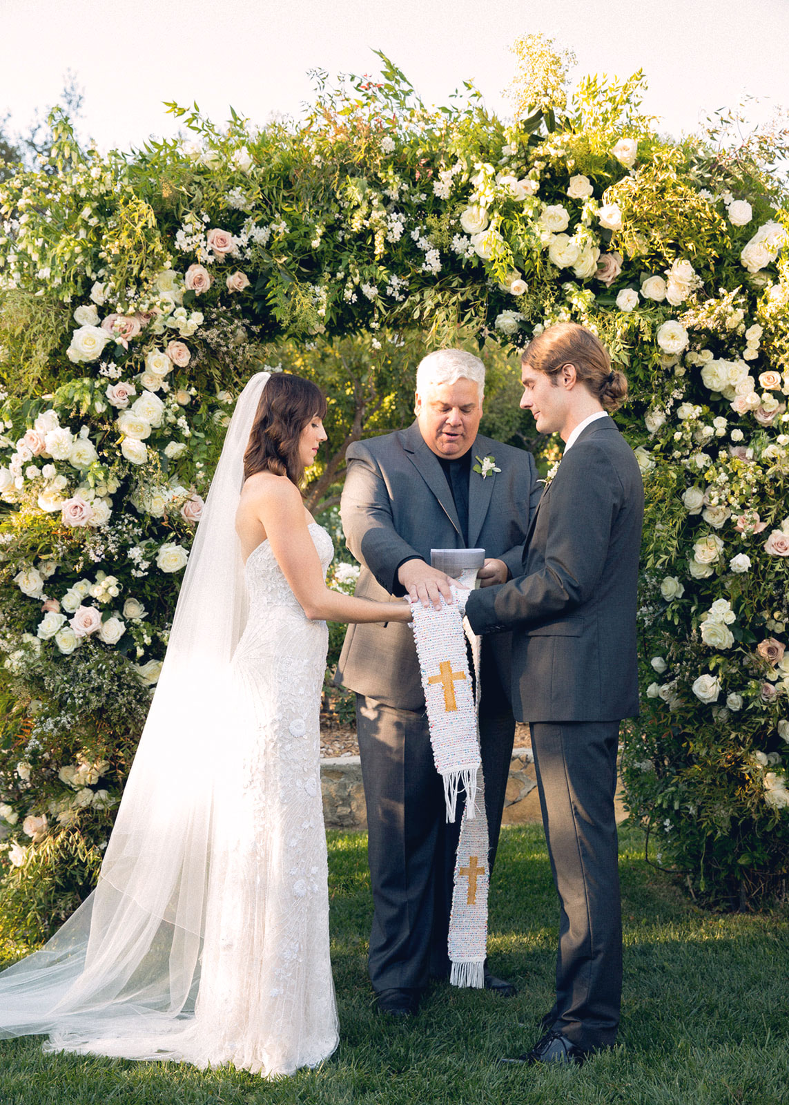 Wide shot of the bride, groom and pastor with their hands all on a bible standing under the flower arch.