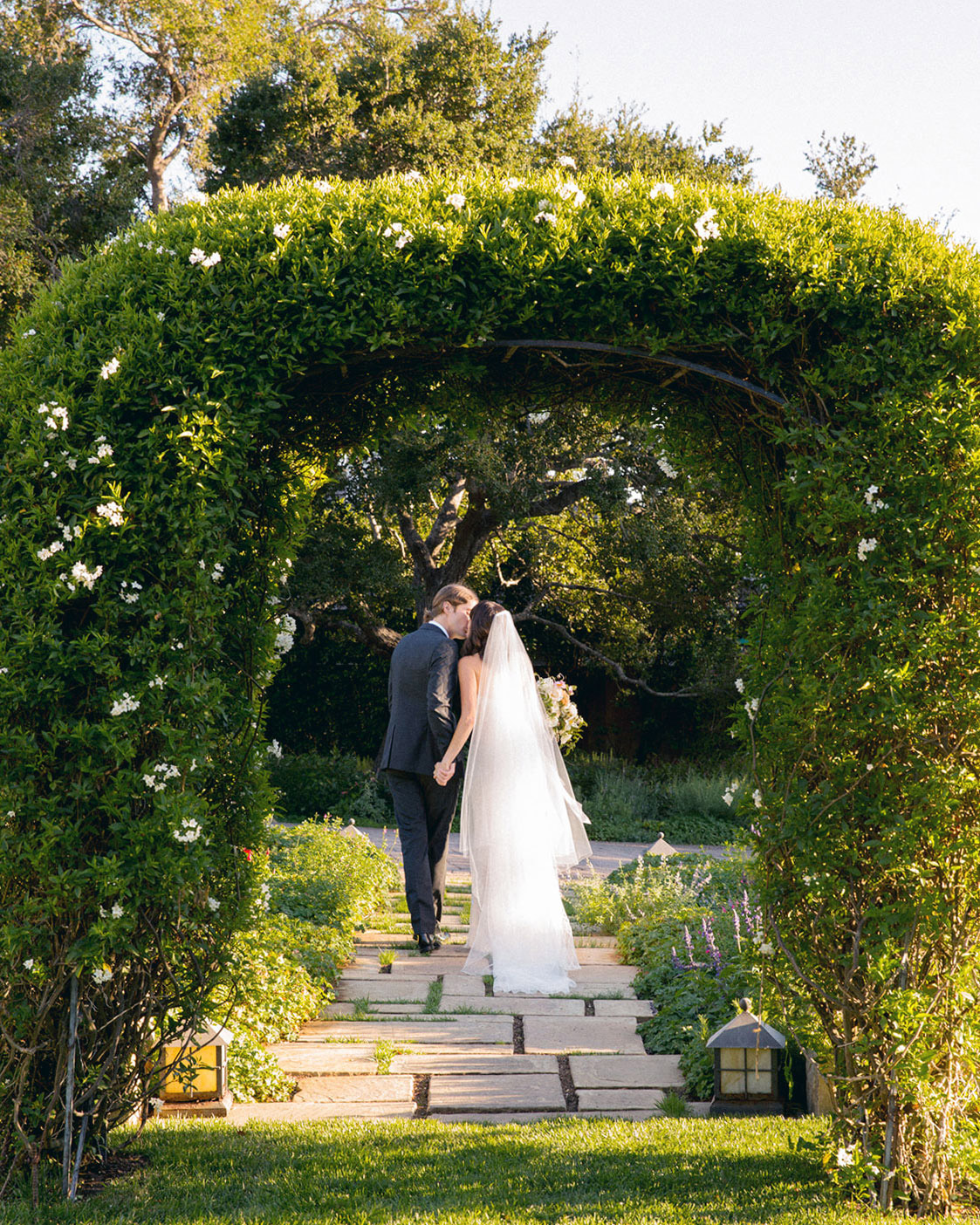 Wide shot of a bride and groom walking away from camera and stopping for a kiss under a green archway.