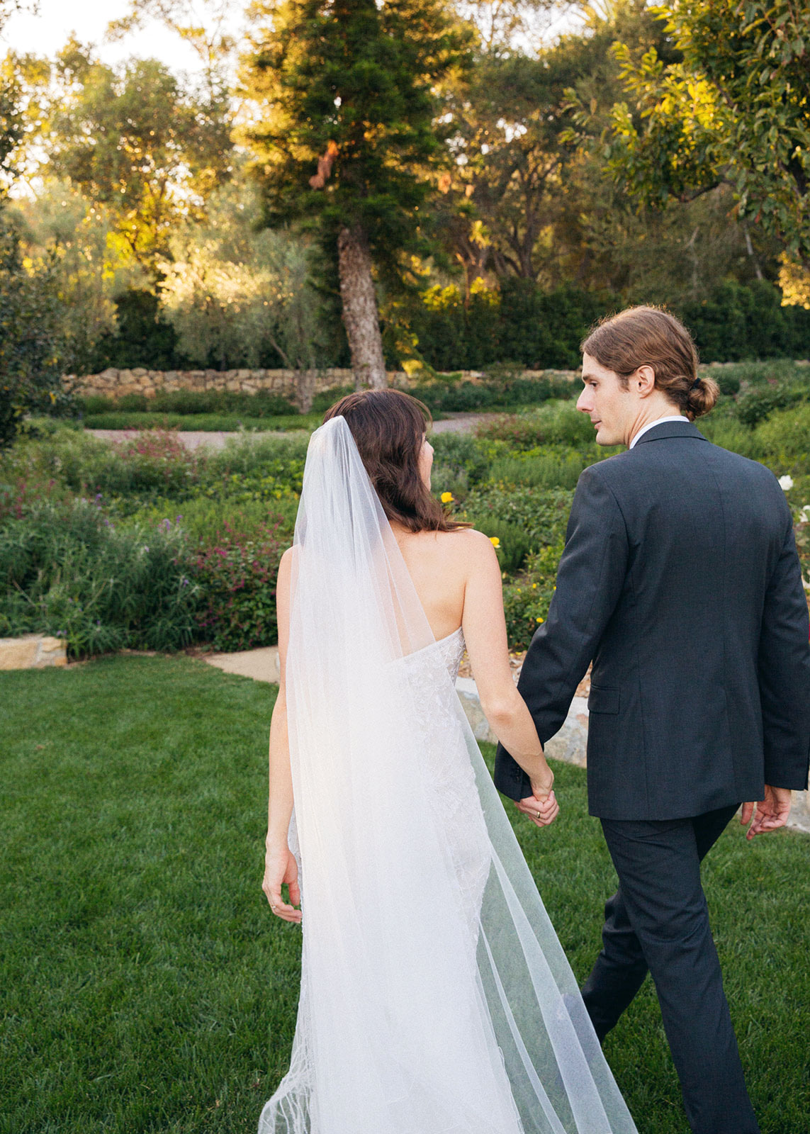 Medium shot of a bride and groom walking away from camera in a big open green garden.