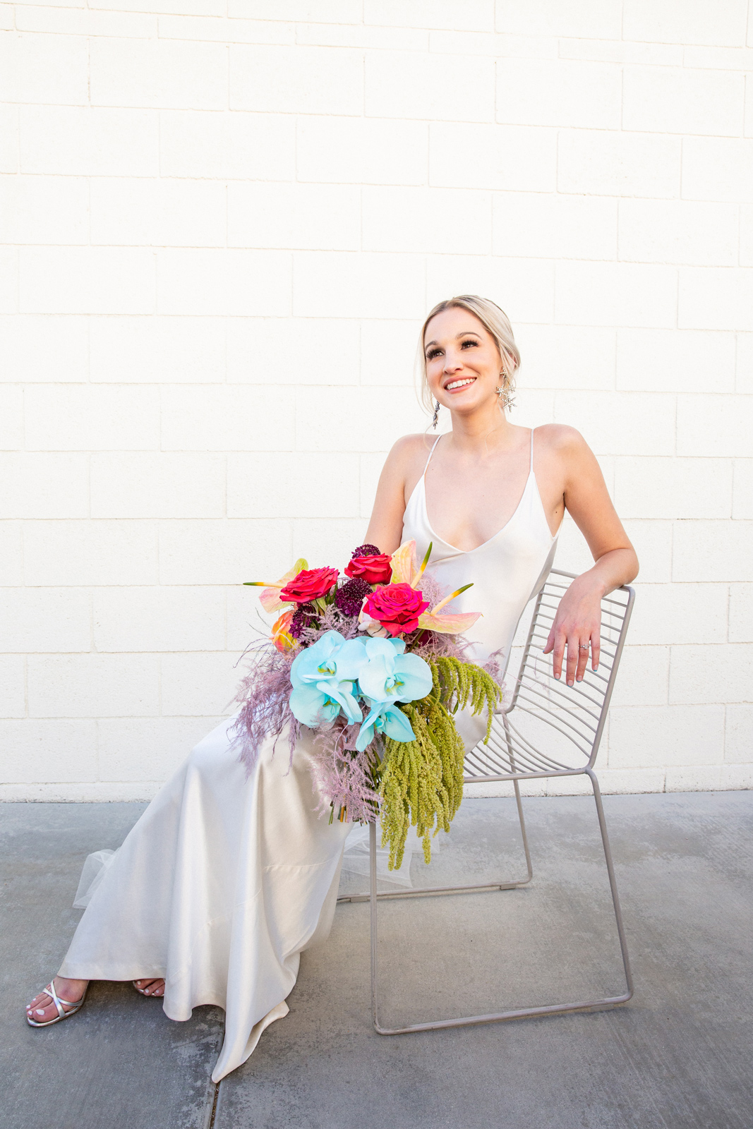Wide portrait of K sitting in her bias cut white silk gown by A la Robe Bridal Paris and Vintage Jimmy Choo shoes on a modern light gray wire geometric sitting chair.