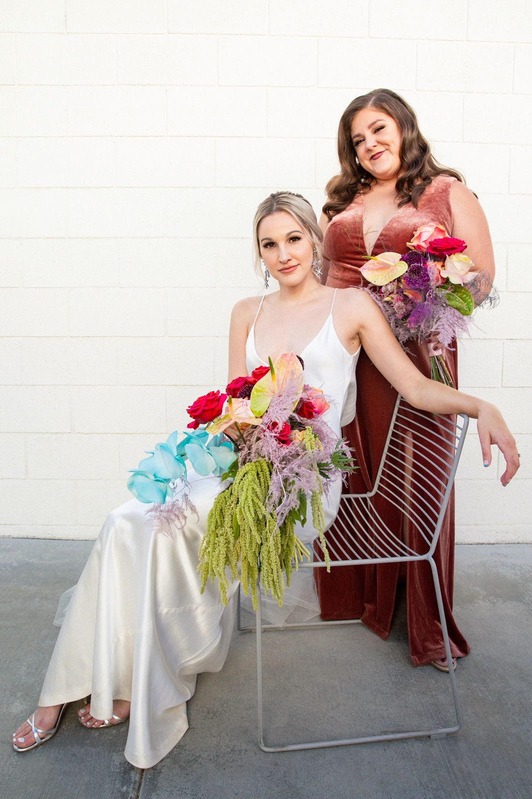 Wide portrait of K sitting in her bias cut white silk gown with her maid of honor wearing a burnt orange velvet full length dress holding a vibrant bouquet of flowers.