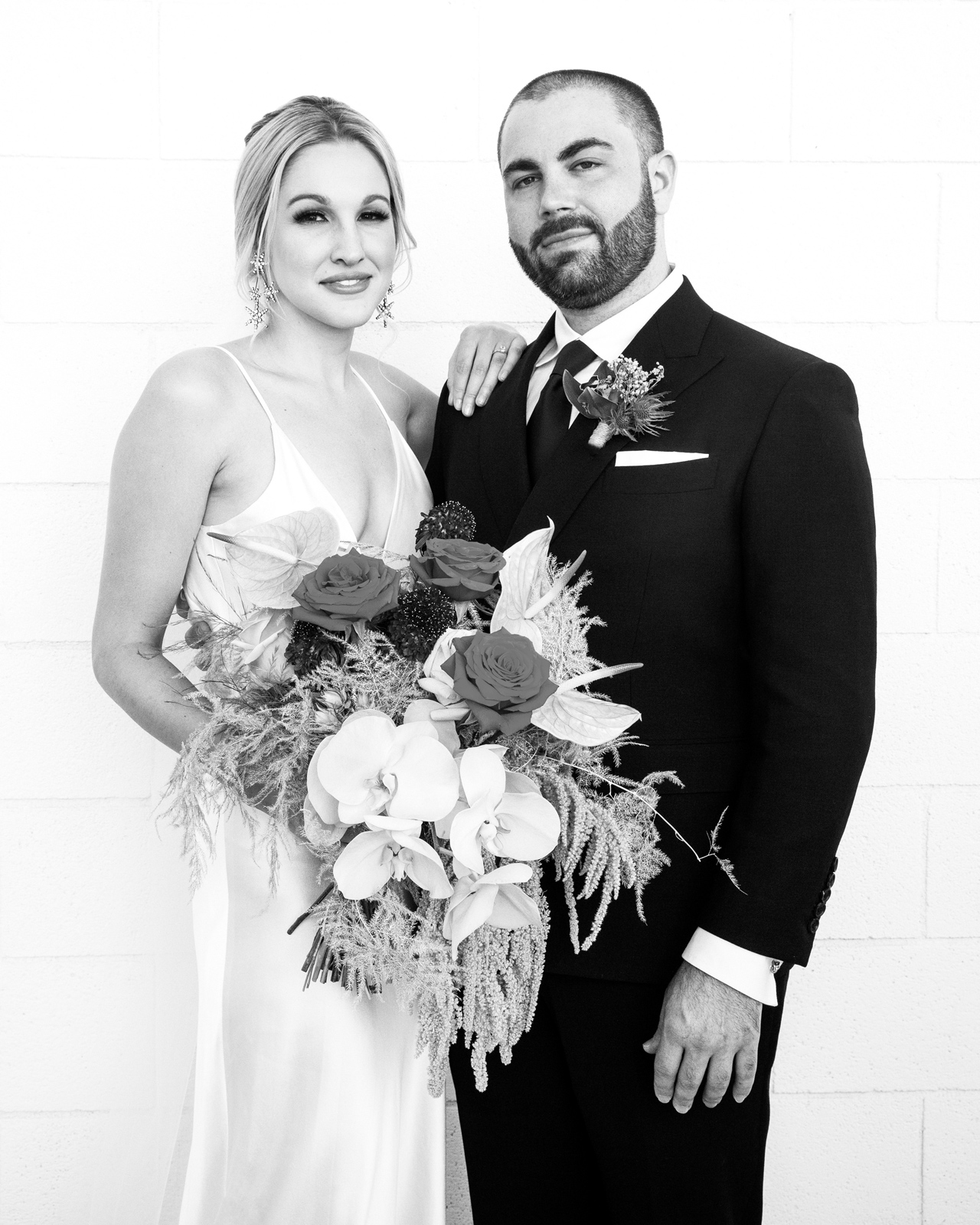 Medium shot of K in her bias cut white silk gown and B in his black double breasted suit jacket tailored by 1710 Bespoke Detroit. This portrait is in black and white with the brides hand on his shoulder and holding her robust wedding bouquet in the other. Both with a small smile looking directly into the cameras eye.
