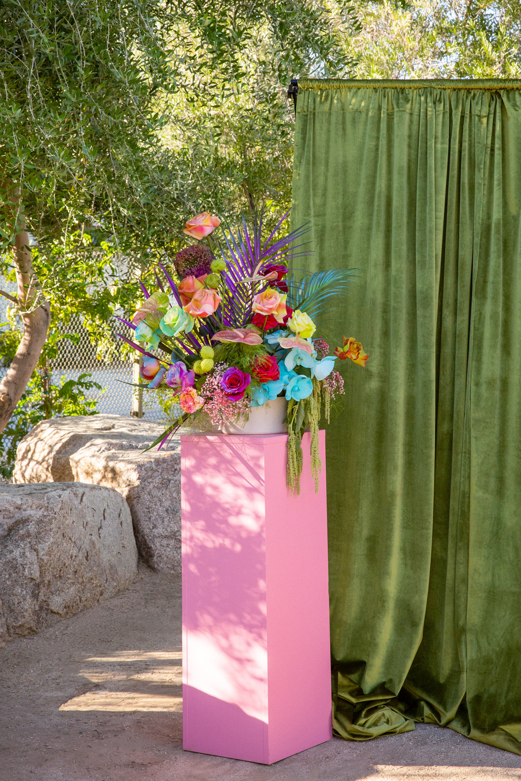Wide shot of a pink pedestal podium holding a large vase of colorful blooms in front of a green velvet curtain.