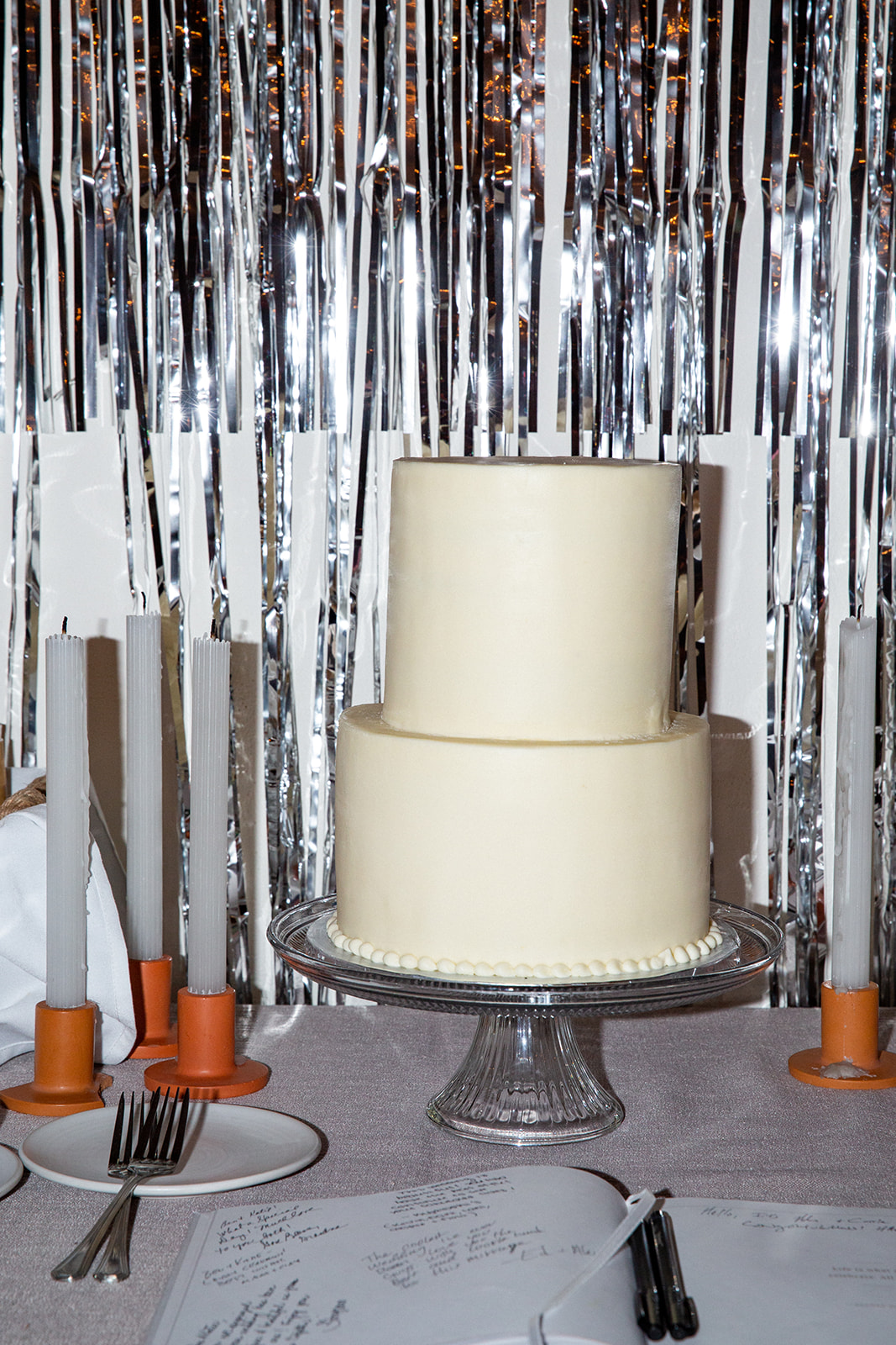 Medium shot of the cake in front of a metallic curtain backdrop. It oversized with 2 layers and is plain off white color and sits on a vintage uranium glass cake stand. K and B also had fun with it by playing “Cake” by Rihanna. during the cutting.
