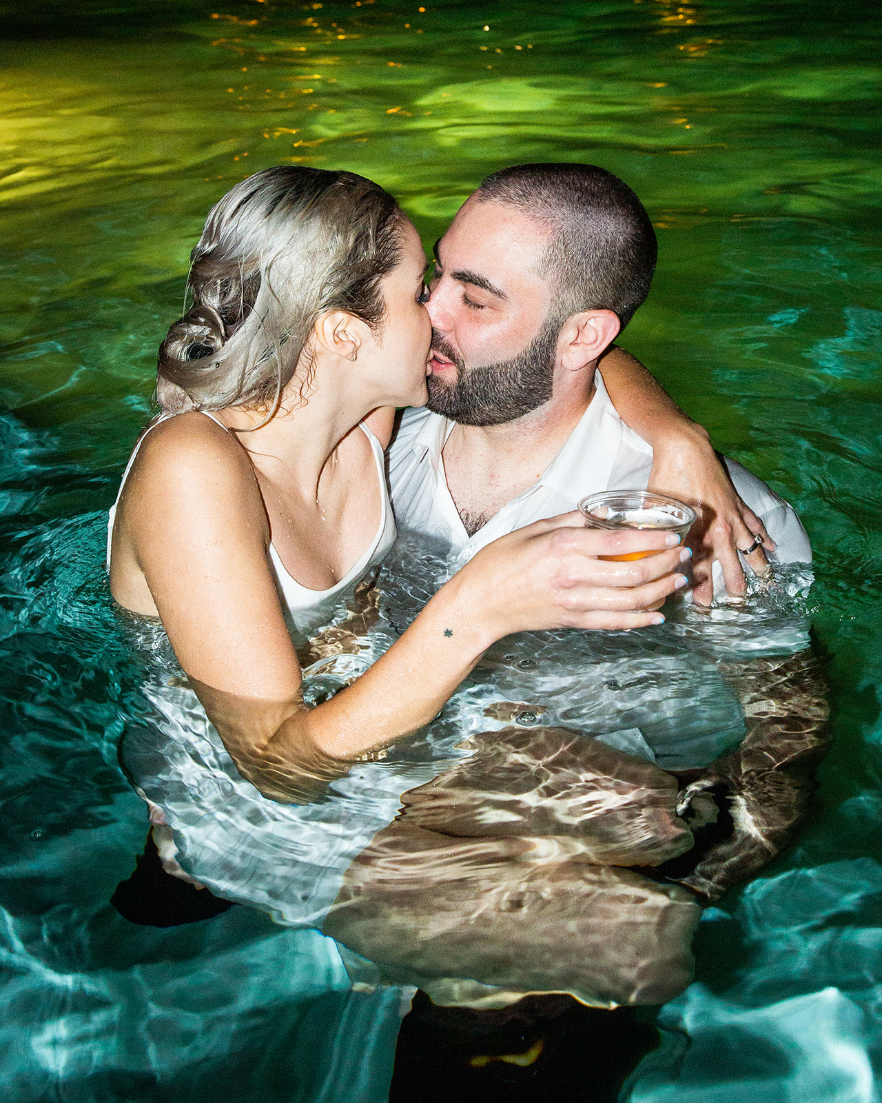Medium shot of K and B in their wedding attire in a swimming pool kissing.