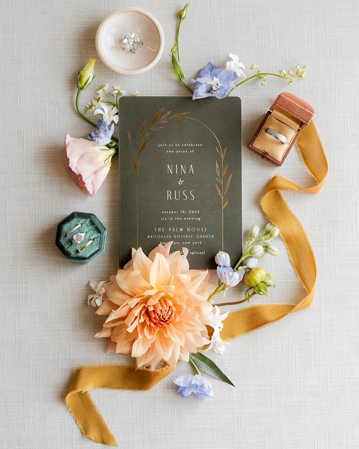 Close-up overhead shot of a wedding invitation surrounded by a large, soft peach-colored dahlia flower, a gold ribbon curling to the right of the frame, and small blue and white flowers scattered around. On the left is a small, velvet green ring box with a diamond engagement ring and two wedding bands. A brown ring box containing another wedding band is placed near the ribbon. At the top, there is a small white dish with sparkling earrings resting inside.