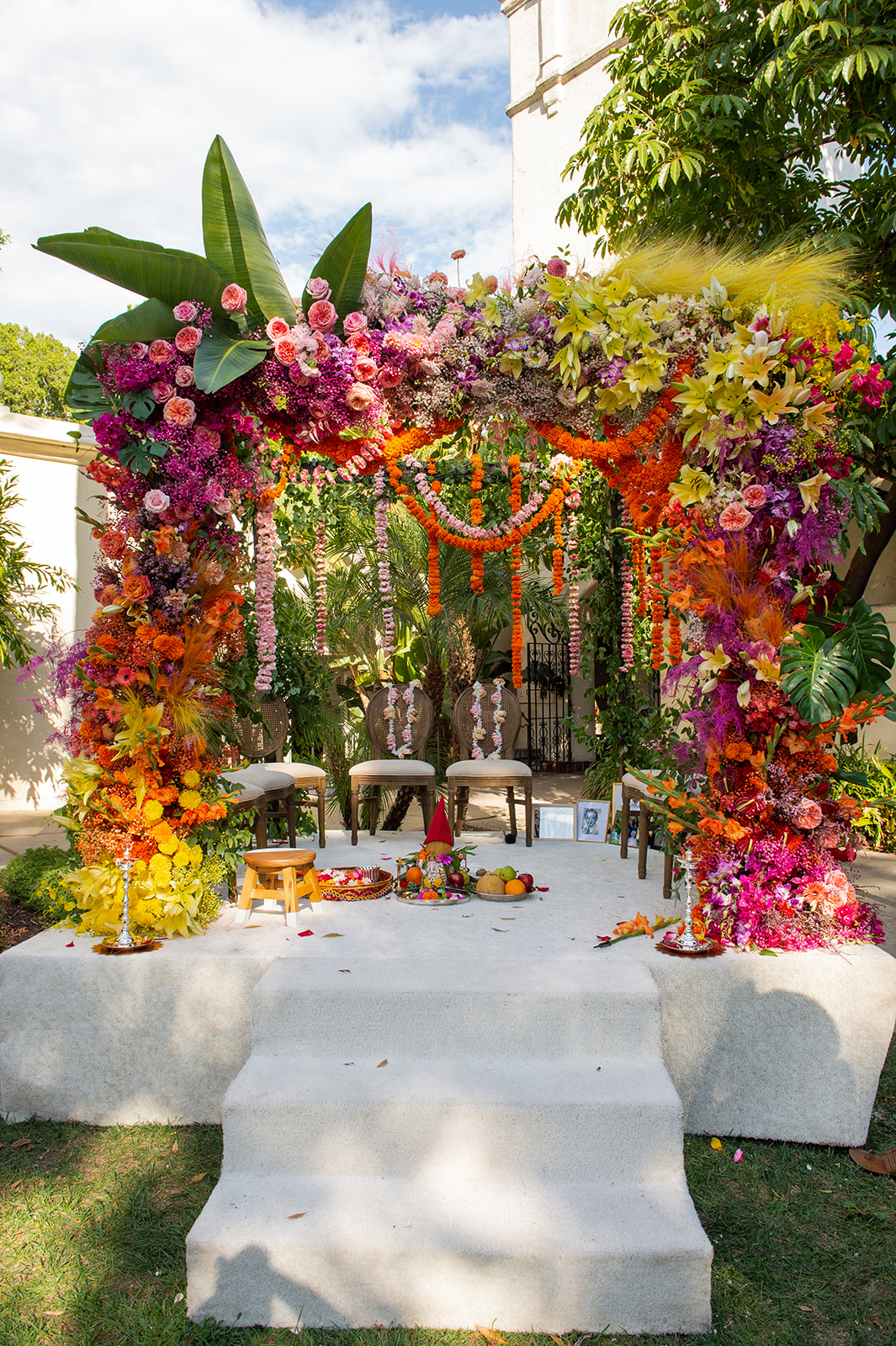 This wide shot depicts a lavishly decorated wedding mandap, a traditional Hindu ceremony setting positioned outdoors under a clear sky. This elaborate structure is adorned with many flowers in a riot of colors ranging from deep purples and pinks to vibrant oranges and yellows. The flowers are arranged in a dense, lush canopy over the mandap, with garlands hanging down, creating a curtain of blooms that frame the central area where the ceremony takes place. Beneath this floral canopy, there are four simple stools for the bride, groom, and officiants, set upon a white, textured fabric that covers the ground. In front of the stools, ritual items are neatly arranged for the ceremony, including fruits, flowers, and a fire pit, essential elements for various Hindu wedding rituals.