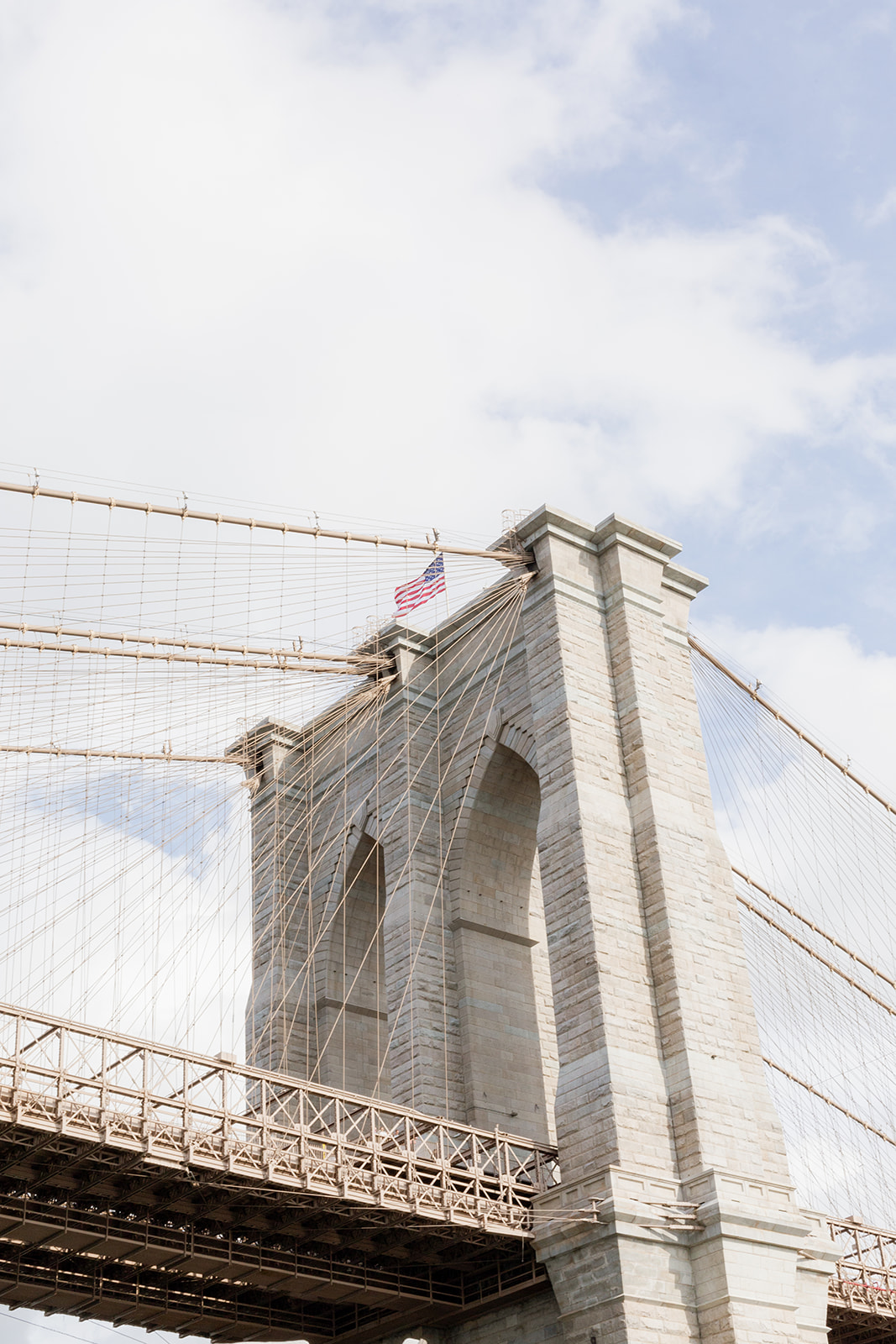 Close-up of a section of Brooklyn Bridge reaching into the light blue sky with soft white clouds scattered across the sky. At the very top of the tower, an American flag flutters in the breeze.