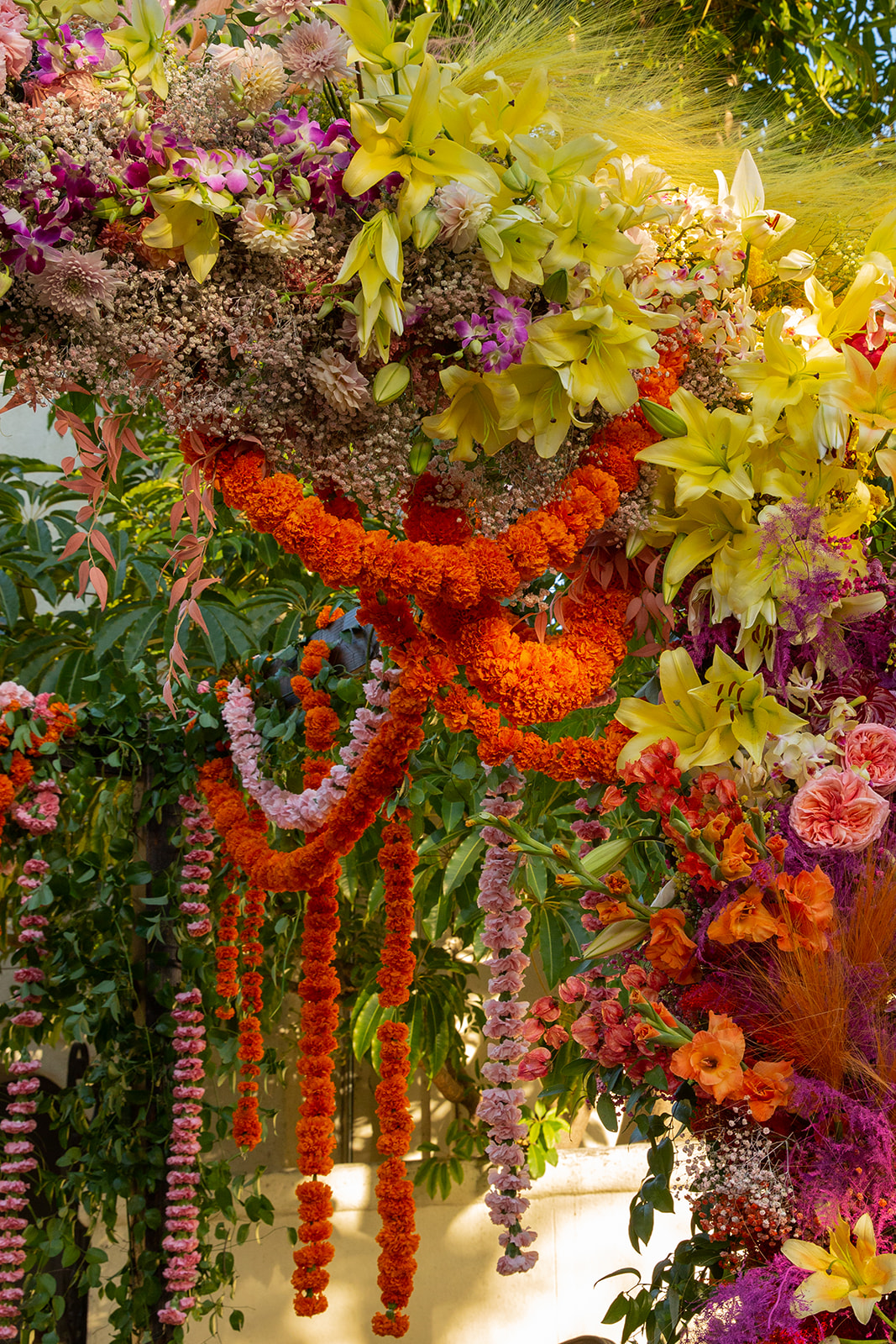 A detailed close-up of floral arrangements on the mandap. Hanging garlands of orange and pink flowers cascade elegantly, drawing the eye with their bold colors and traditional styling. These garlands create a visual pathway that frames the rest of the floral arrangement, enhancing the setting