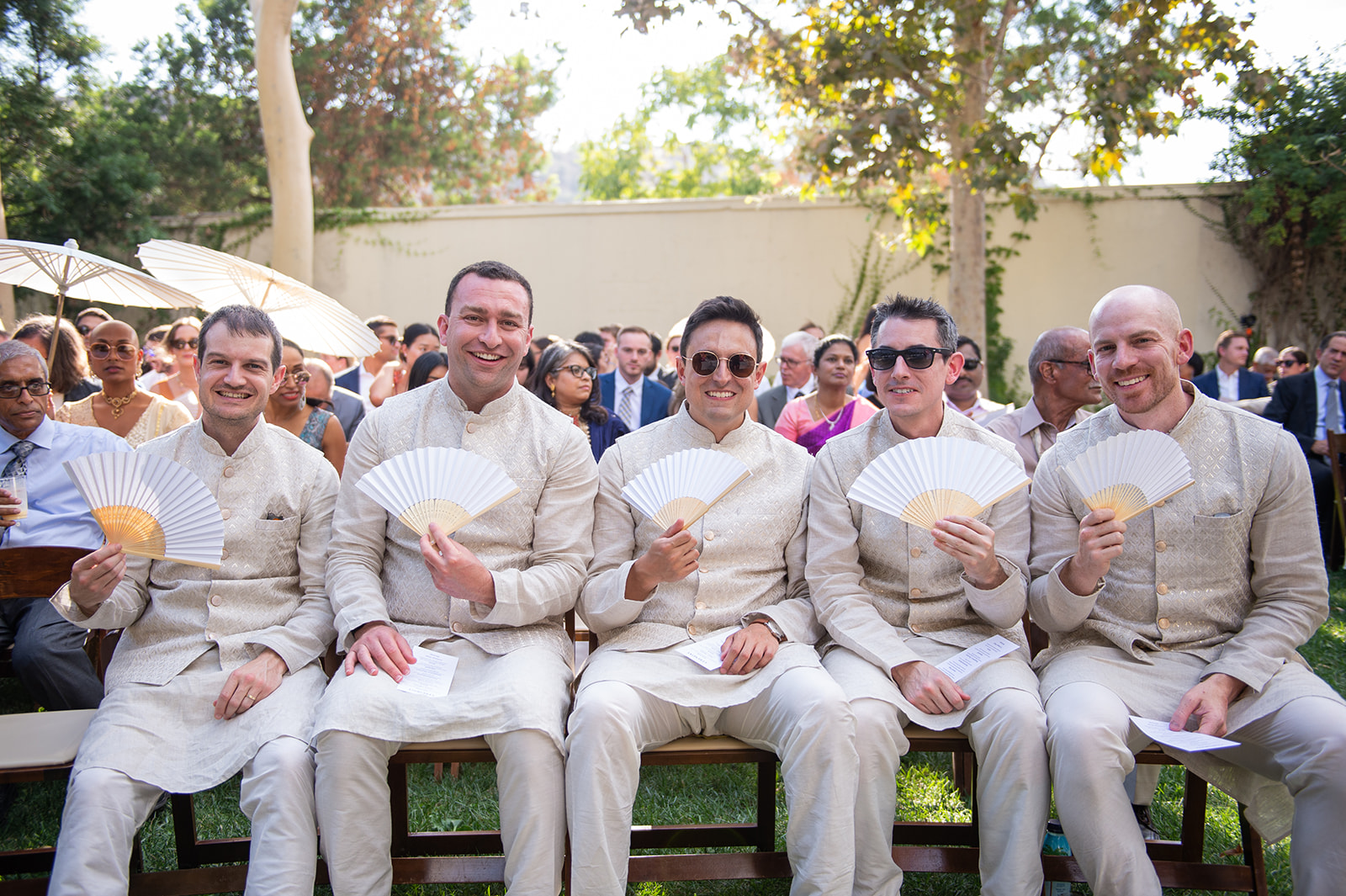 Wide shot of a group of 5 groomsmen smiling and looking into the camera. They are dressed uniformly in traditional cream-colored Indian outfits, which include intricate embroidery on the fabric. The men are holding delicate white fans to keep cool from the heat.