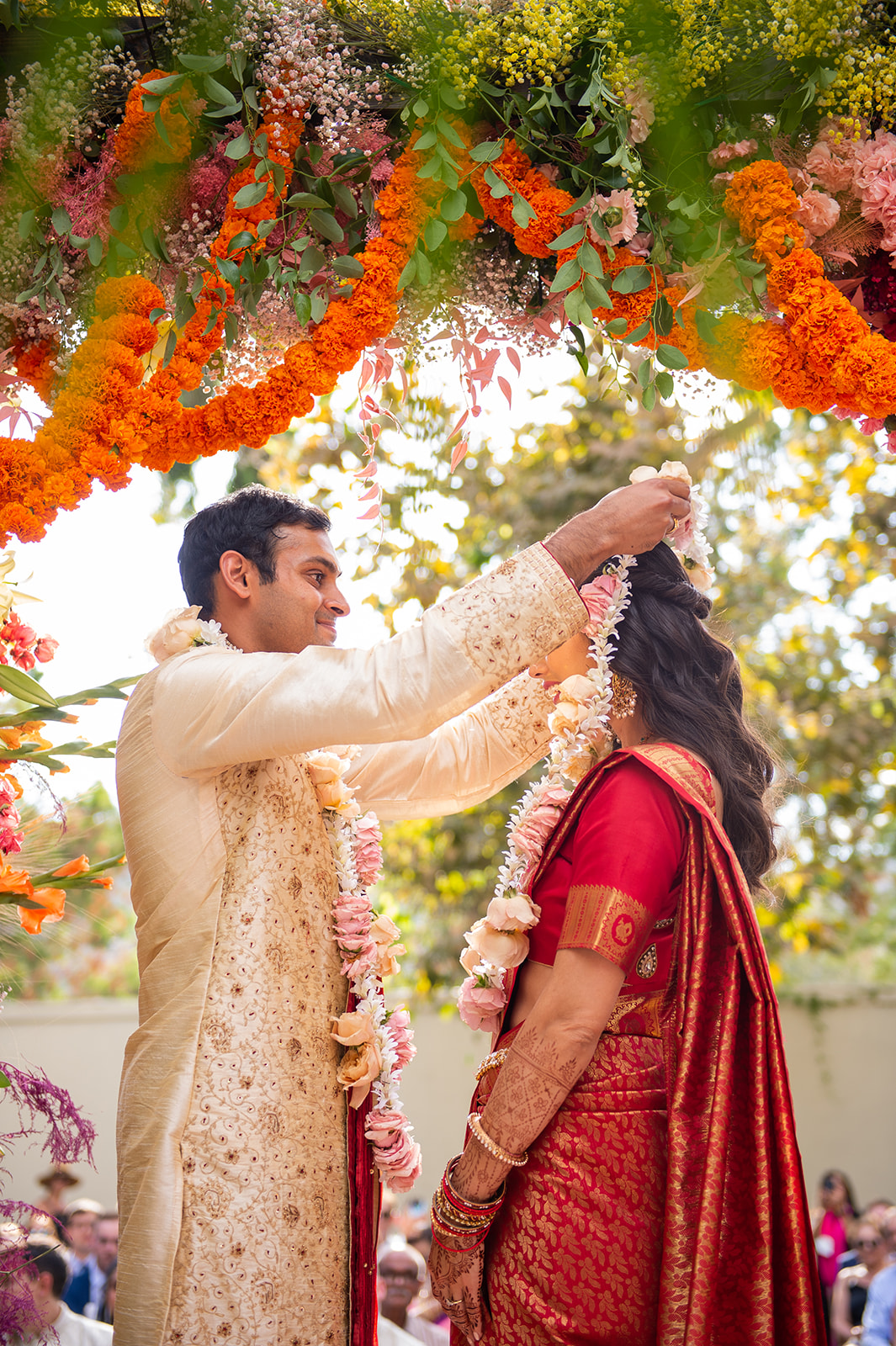 Capture of bride and groom standing under a lavishly decorated floral arch of the mandap, vibrant with colors ranging from orange to pink to purple. The groom, dressed in an ornate cream sherwani, is seen placing a garland of white flowers around the bride