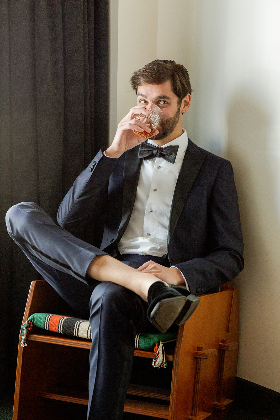Medium close-up portrait of our groom, R, sitting casually in a chair. He is dressed in a dark, well-fitted tuxedo with a black bow tie, white dress shirt, and black velvet loafers. Sipping from a glass with an amber-colored drink. His leg is crossed, with one foot resting on his knee.