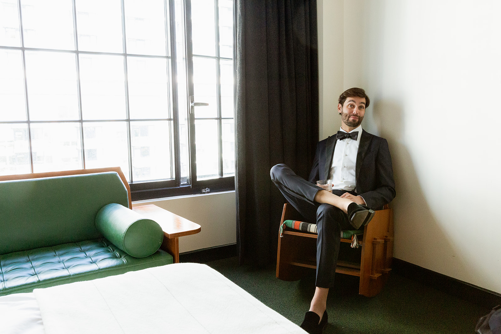 A wide shot of our groom, R, sitting in a hotel room is softly lit by large, bright windows framed by black curtains to the left. Large windows let in a lot of natural light, illuminating the room.