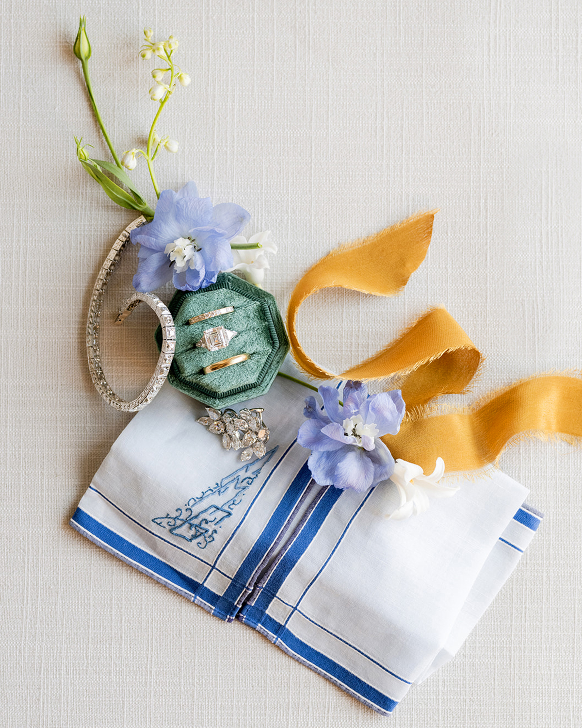 Close-up shot of an overhead, flat-lay photograph of a collection of wedding day items. At the center is a soft, green, velvet ring box with three rings, including a diamond engagement ring and two wedding bands. Surrounding the box is a sparkling diamond bracelet, a pair of earrings with intricate stones, and a white handkerchief with blue embroidered details. A delicate marigold-colored ribbon flows across the right side of the image, adding a soft contrast. Small flowers are scattered throughout the arrangement, including a light purple bloom and tiny white buds.