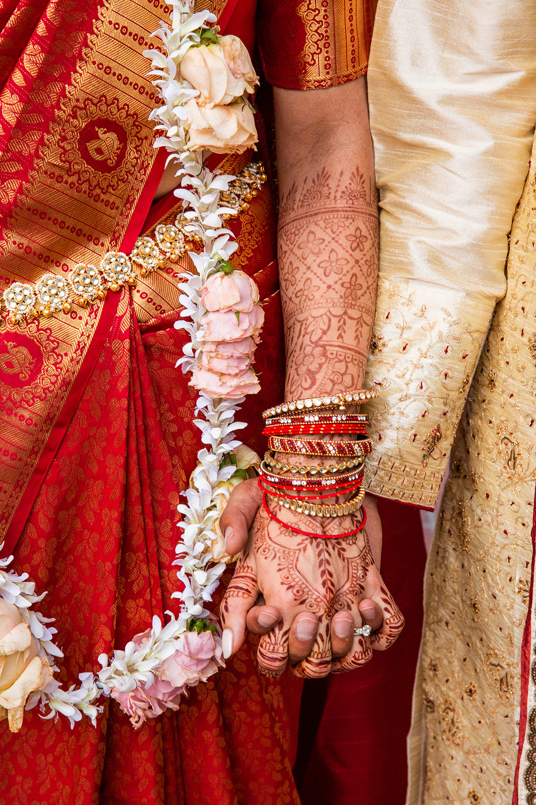 Close-up shot of the intricate details of the bride’s hands are adorned with detailed mehndi (henna), which signifies beauty, joy, and spiritual awakening. She wears ornate red bangles and holds a string of flowers, which are often symbols of good luck and prosperity. The groom’s hand is partially visible, hinting at his traditional attire that complements the bride’s vibrant red saree adorned with golden embroidery.