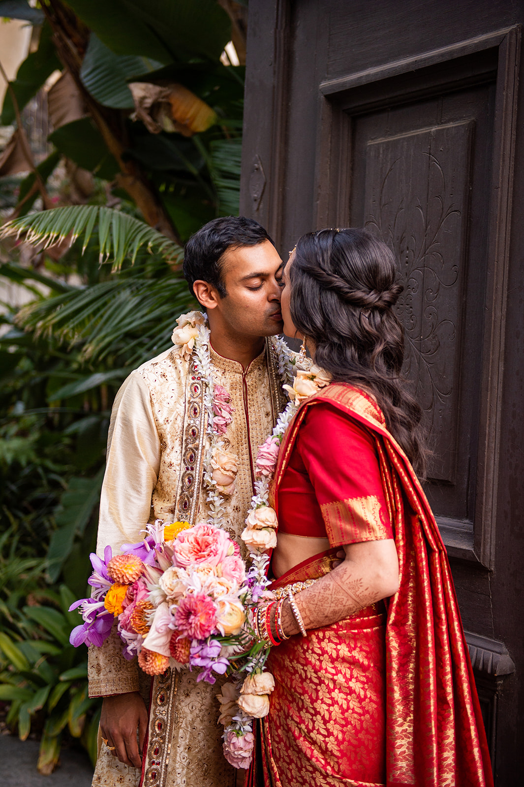 Medium shot of the couple kissing in front of a large wooded door with a lush green garden scene behind them.