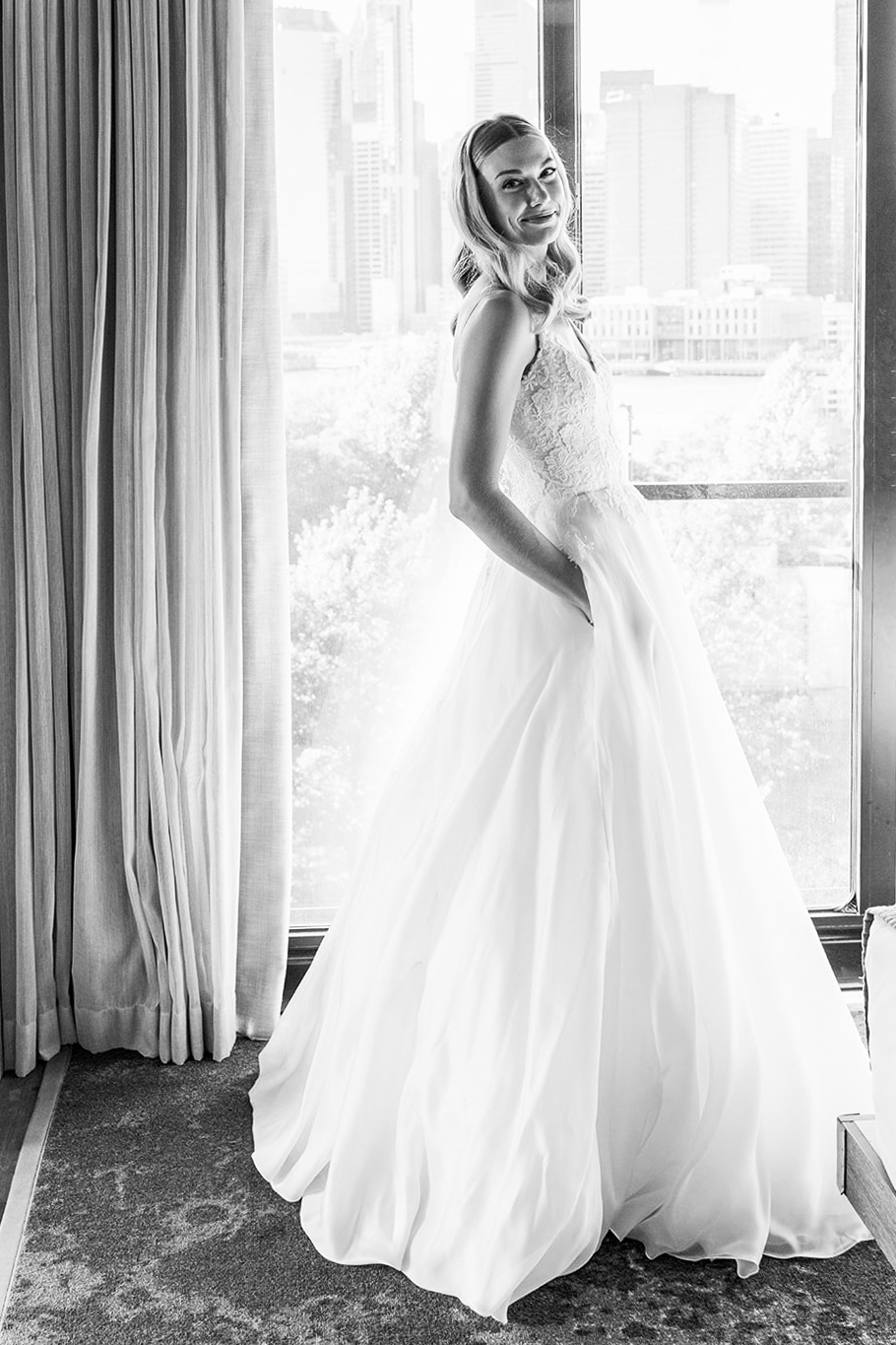 This black-and-white photograph shows our bride, N, posing gracefully in front of a large window. She is wearing a classic, elegant floor-length wedding gown designed by Caroline Castigliano with a fitted lace bodice and a full, flowing skirt that billows softly around her.