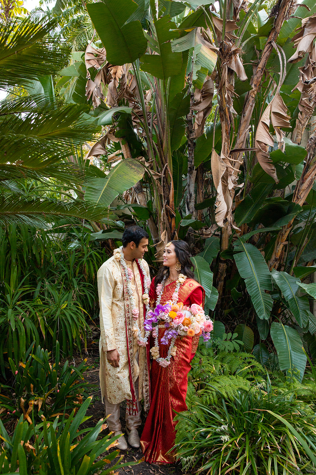 Fullbody portrait of our couple in the lush green setting looking at each other and smiling.