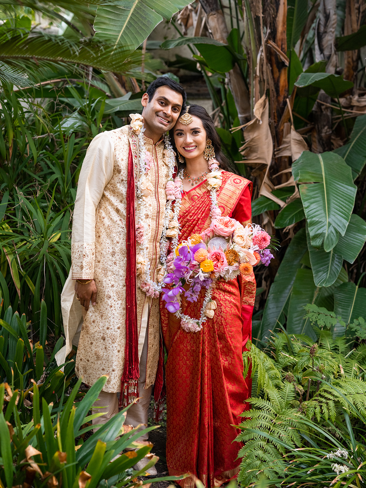 Fullbody portrait of our bride and groom in a lush garden setting, reflecting the joy and beauty of their wedding day. The bouquet she holds adds a burst of color with its mixture of flowers in shades of pink, orange, purple, and yellow, blending beautifully with the natural greenery surrounding them